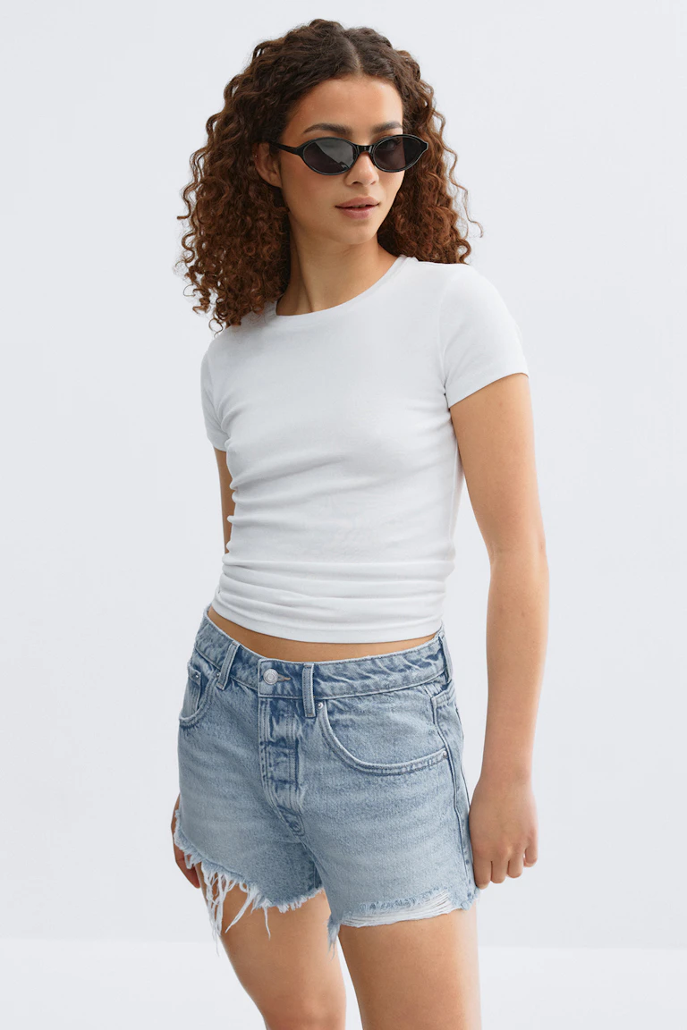 Witte top met korte mouwen, lichtgekleurde denim shorts met hoge taille en gerafelde zoom en een zwarte zonnebril met kattenogen.