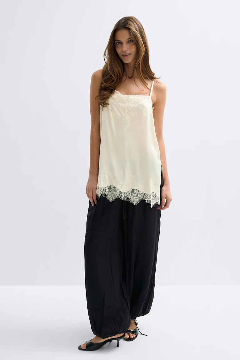 Camisole en soie crème à fines bretelles bordée de dentelle, associée à un pantalon ample noir à cordon de serrage et à des sandales noires à talons aiguilles.