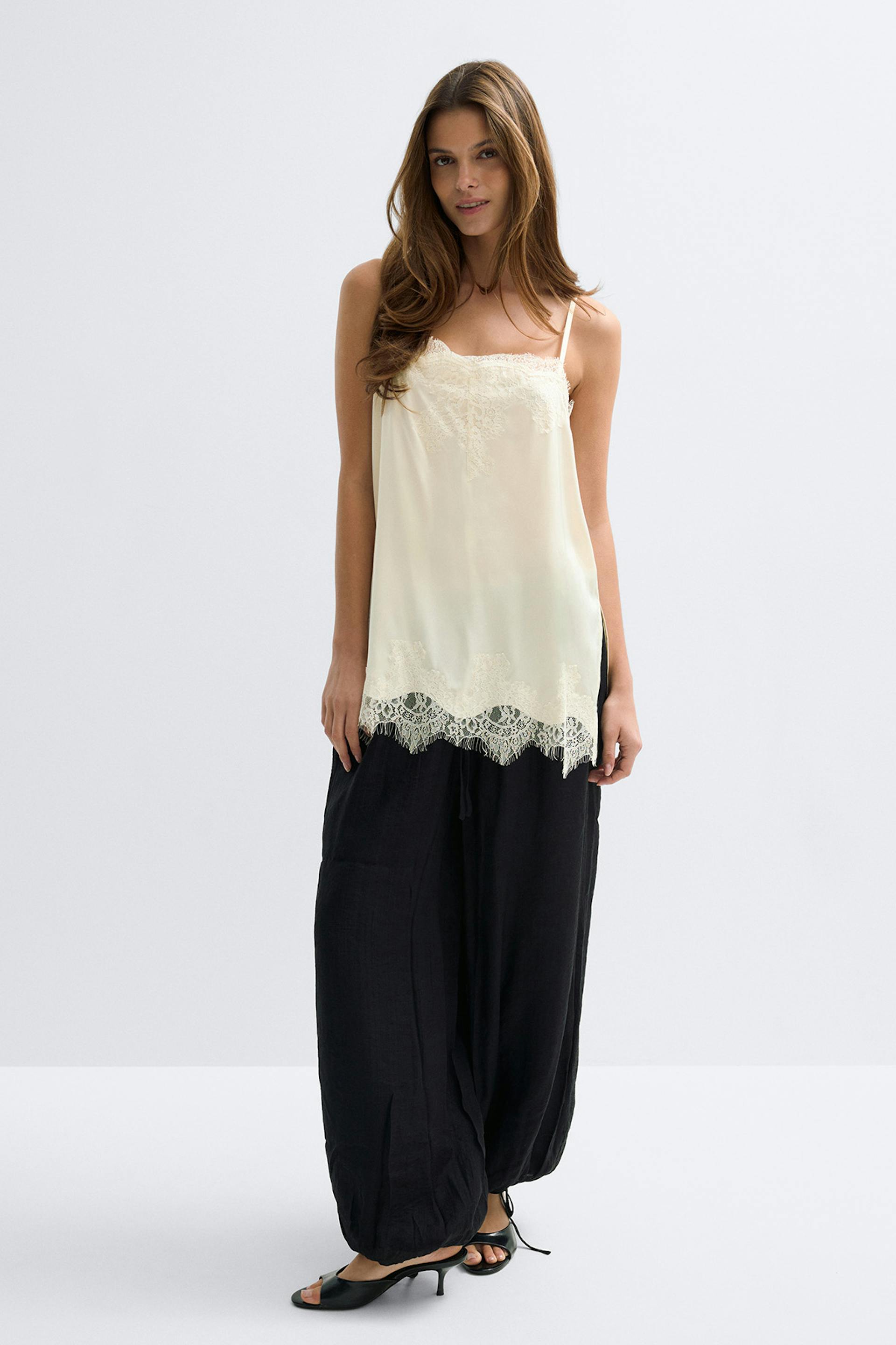 Camisole en soie crème à fines bretelles bordée de dentelle, associée à un pantalon ample noir à cordon de serrage et à des sandales noires à talons aiguilles.