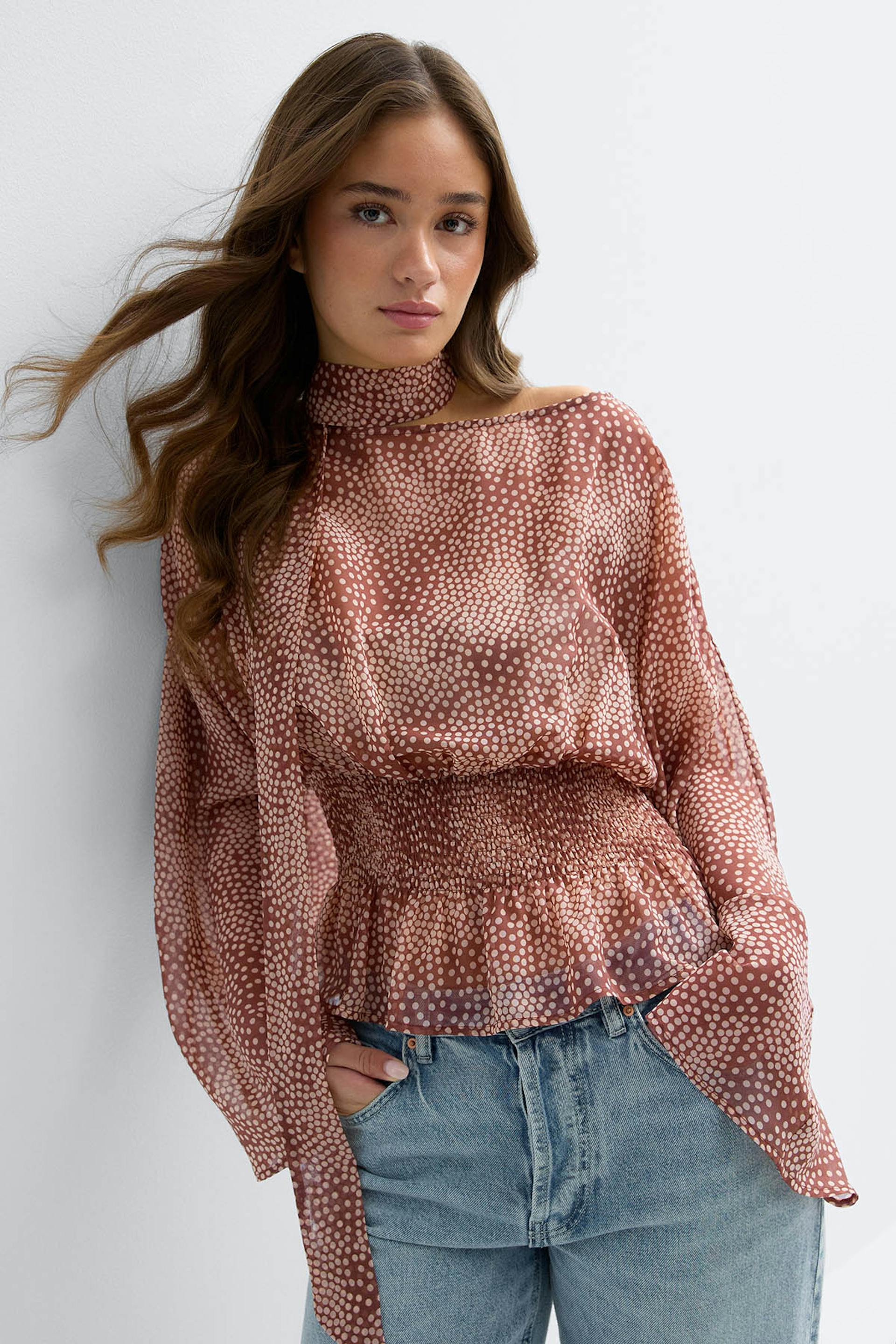 Woman wearing a rust-colored blouse with white polka dots — high tie-neck, long billowy sleeves, smocked waist and peplum hem — paired with light-wash blue jeans.