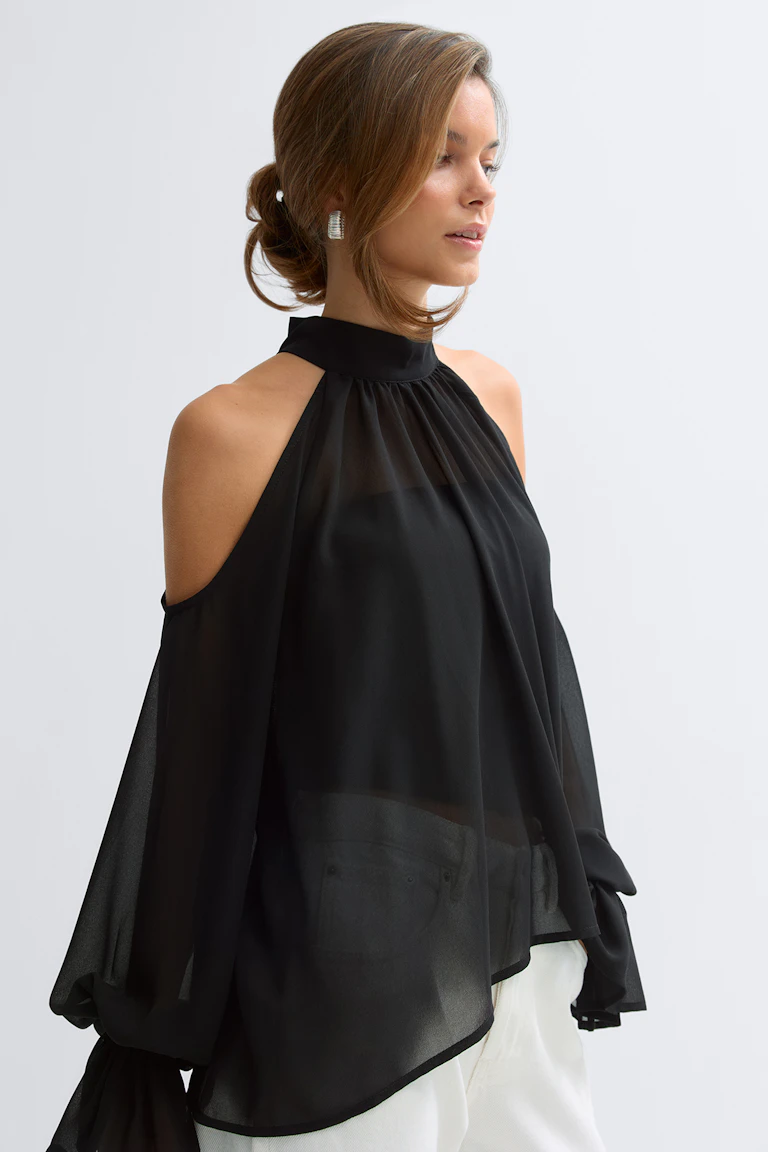 Schwarze, transparente, schulterfreie Bluse mit hohem Ausschnitt und drapierten Läuferärmeln mit gebundenen Manschetten, getragen zu einer weißen Hose.