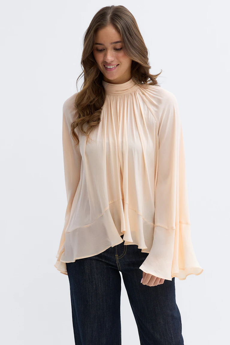 Pfirsichfarbene Chiffonbluse mit hohem Halsausschnitt, geraffter Passe, mehrlagig drapiertem Saum und ausgestellten Ärmeln, kombiniert mit dunklen Denim-Jeans.