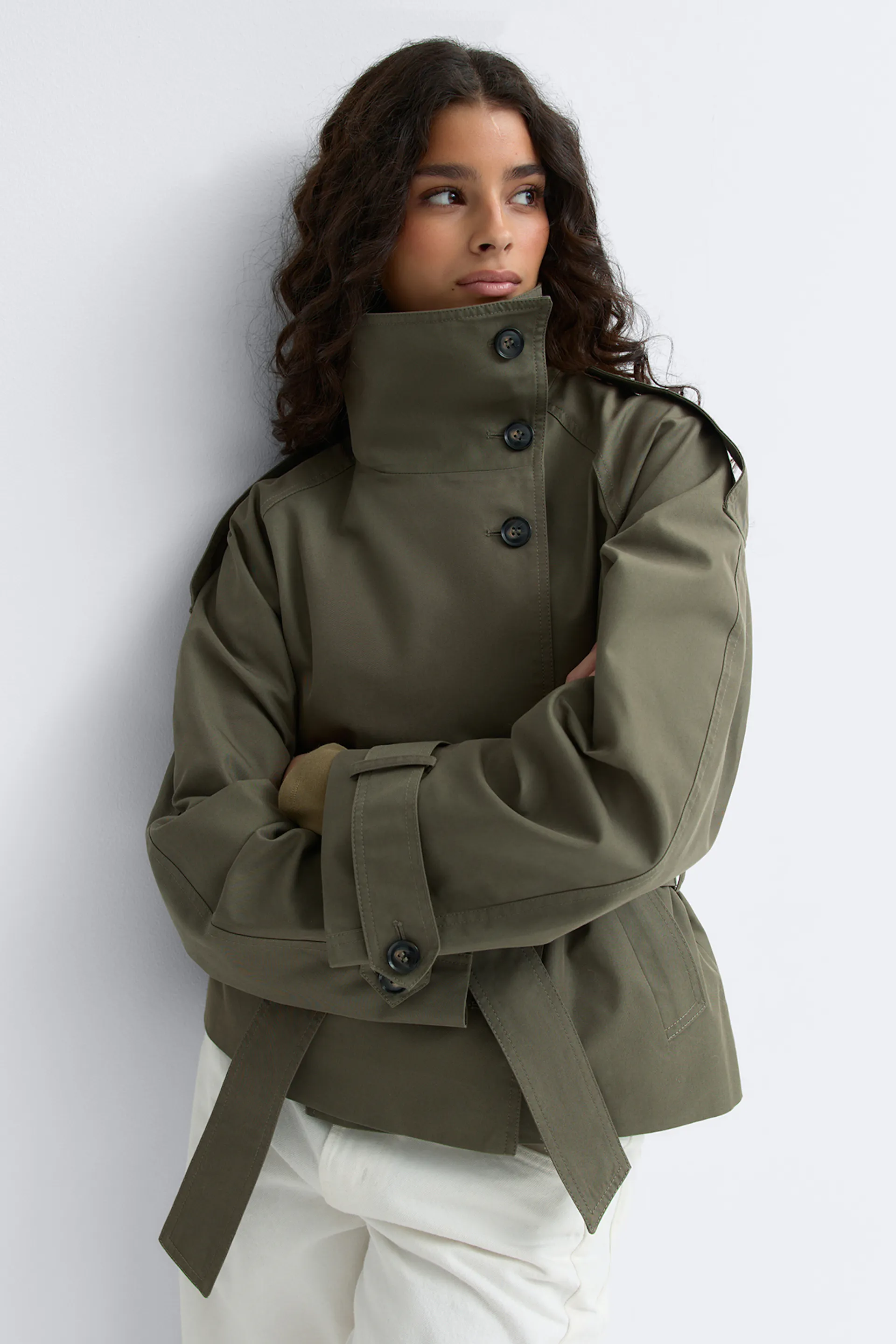 Olivgrüner Trenchcoat mit hohem, geknöpftem Stehkragen, Schulterklappen, verstellbaren Gürtelmanschetten und Taillengürtel, kombiniert mit einer weißen Hose.