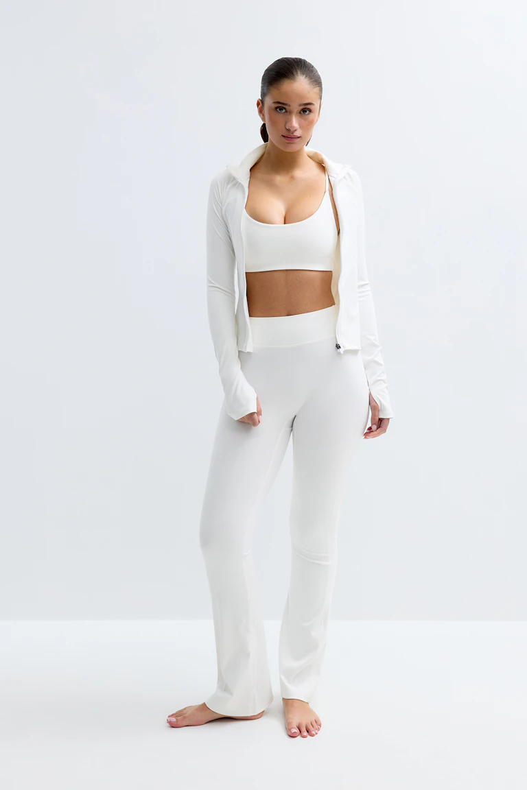 Hvidt sæt med aktivt tøj: sports-bh med rund hals, langærmet jakke med hætte og lynlås med huller til tommelfingrene og højtaljede, udspilede leggings; barfodet.
