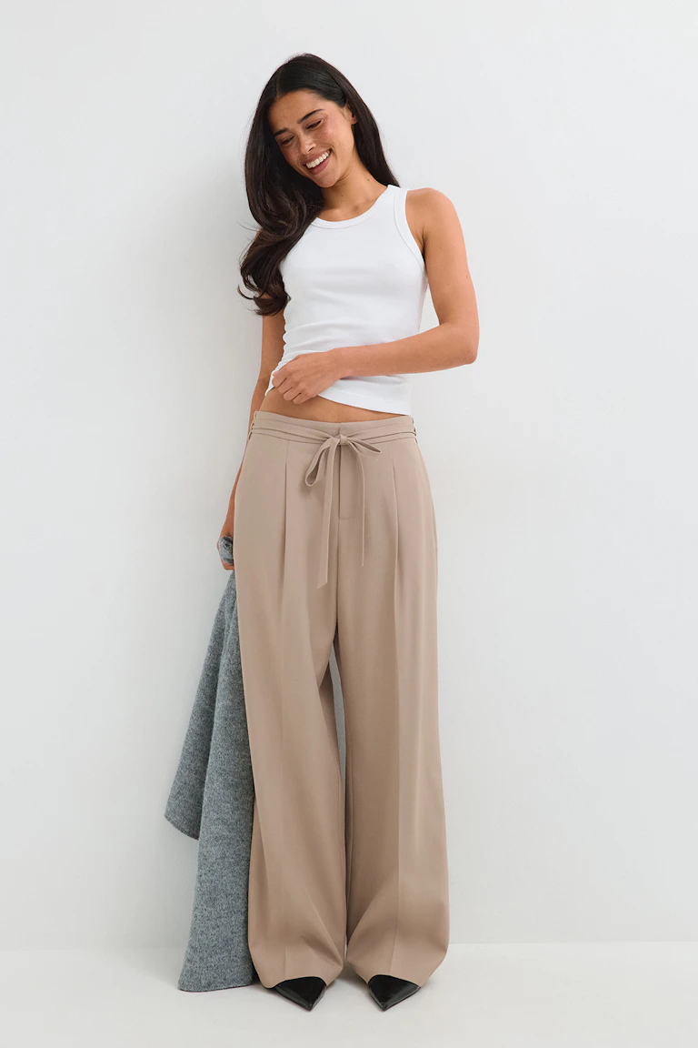 White sleeveless top, beige wide-leg trousers, black pointed-toe shoes, and a grey coat draped over one arm.
