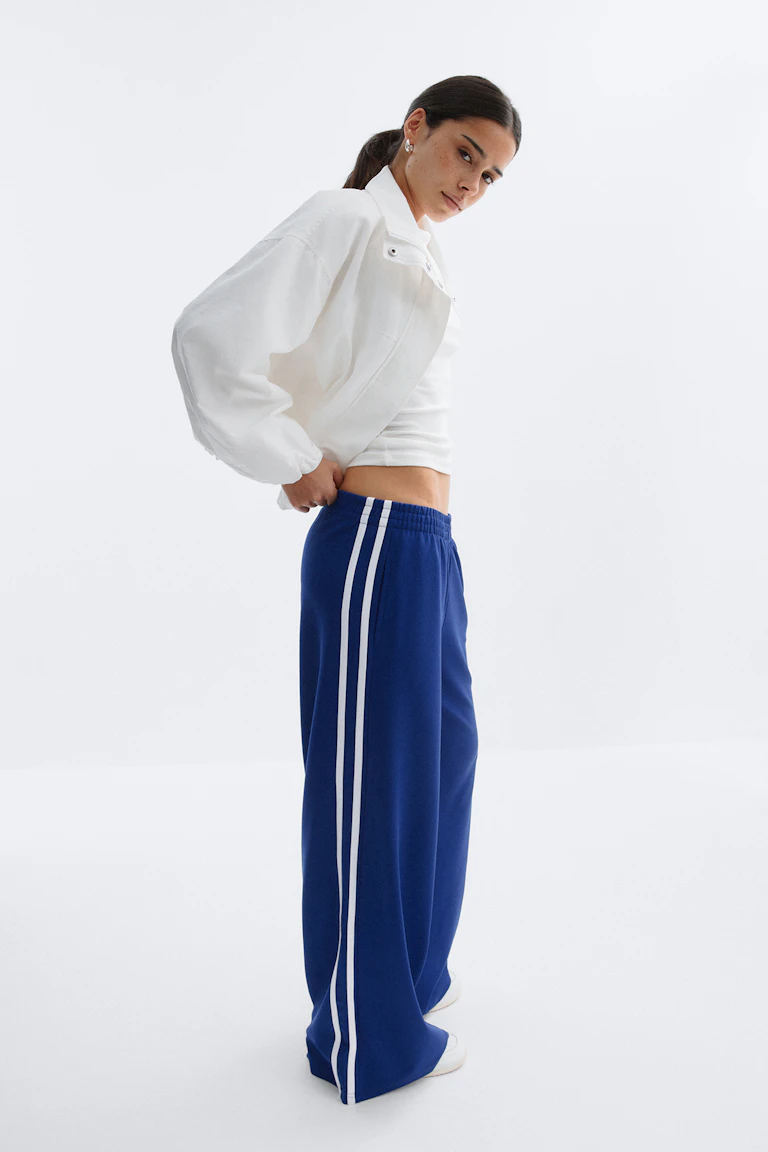 Vit croppad jacka med dragkedja över ett vitt croppat linne, kombinerat med koboltblå vida träningsbyxor med dubbla vita sidoränder och elastisk midja samt vita sneakers.