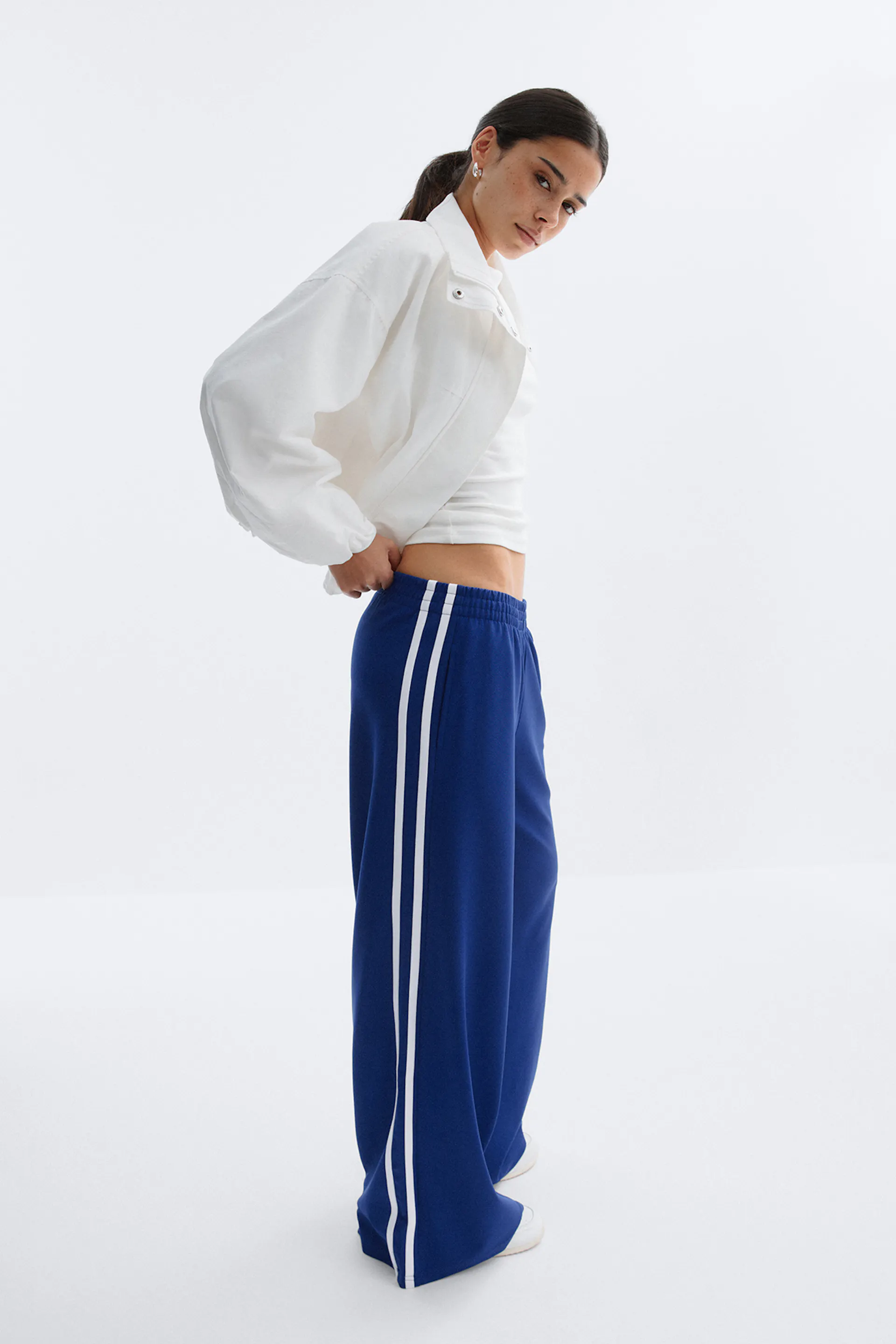 Hvid, cropped jakke med lynlås over en hvid, cropped tank, sammen med koboltblå, brede træningsbukser med to hvide sidestriber og en elastisk linning og hvide sneakers.