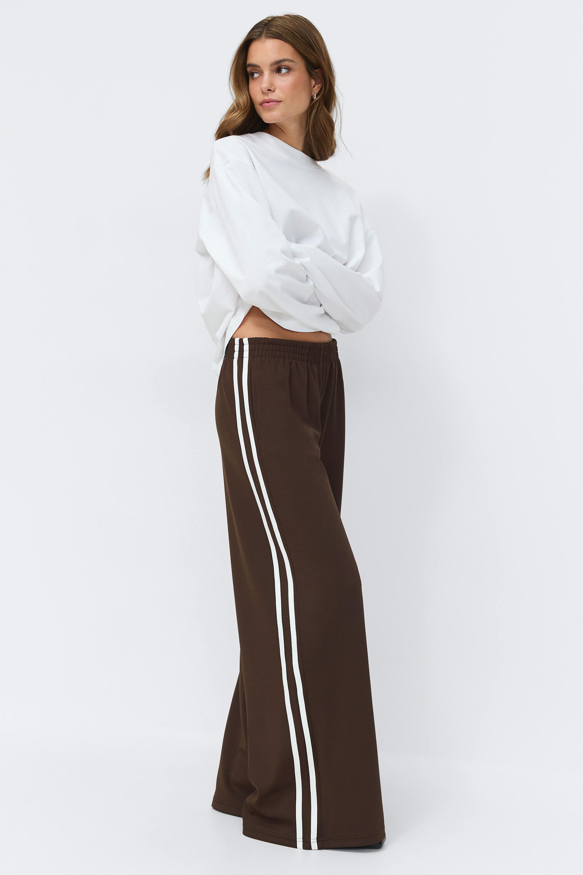 White oversized long-sleeve sweatshirt paired with brown wide-leg track pants featuring twin white side stripes and an elastic waistband.