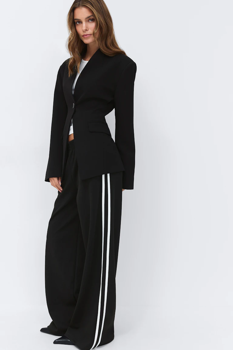 Schwarzer maßgeschneiderter Blazer mit einem einzigen Knopf über einem weißen Oberteil mit Rundhalsausschnitt, kombiniert mit einer schwarzen Trainingshose mit weitem Bein und zwei weißen Seitenstreifen und spitzen schwarzen Schuhen.