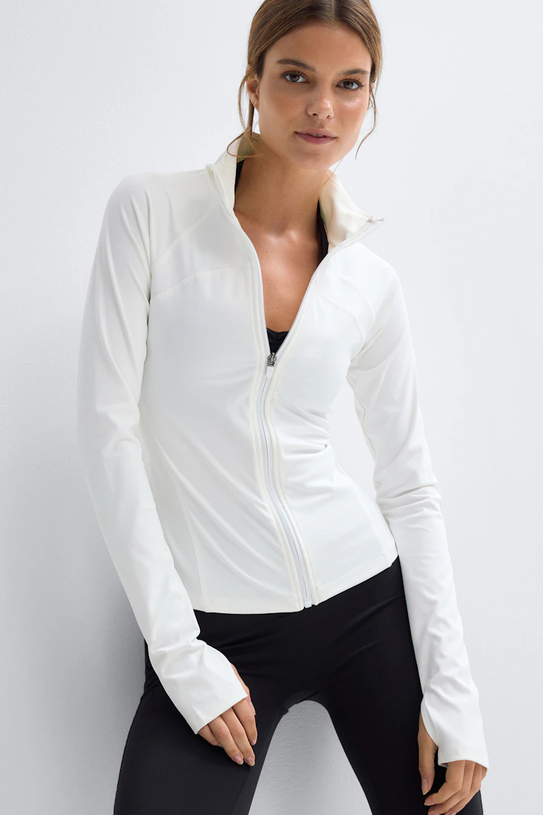 Vit långärmad jacka med hel dragkedja, hög krage och tumhål, över en svart sport-bh och tillsammans med svarta leggings.