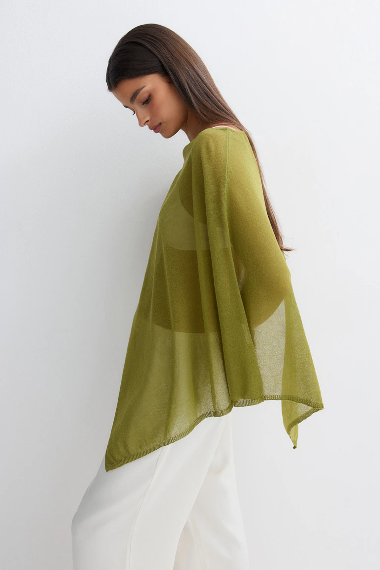 Half doorschijnende olijfgroene gebreide poncho-top in combinatie met een witte broek met wijde pijpen.