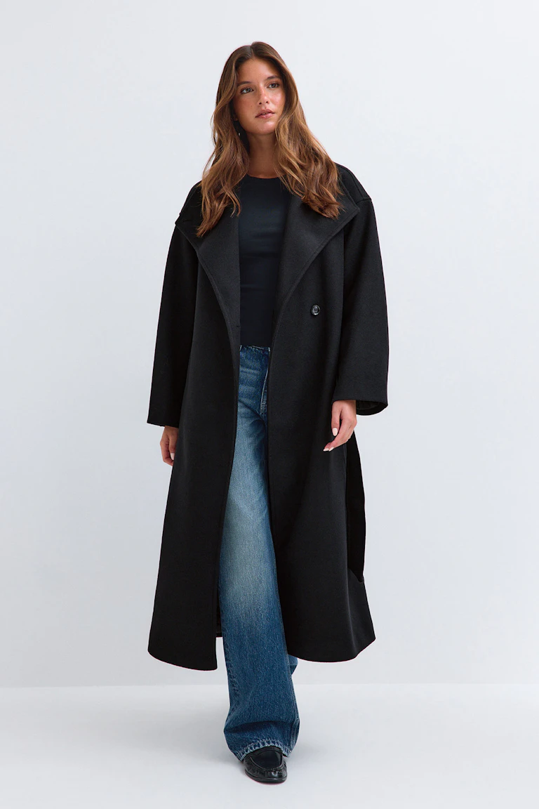 Ein langer schwarzer Mantel über einem schwarzen Oberteil, dazu eine blaue Jeans und schwarze Slipper.