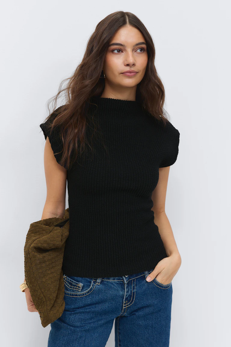 Svart kortärmad topp med mockahals i ribbstickat material tillsammans med mellanblå jeans med kontrastsömmar, kompletterat med ett guldarmband och en olivfärgad vävd clutch.