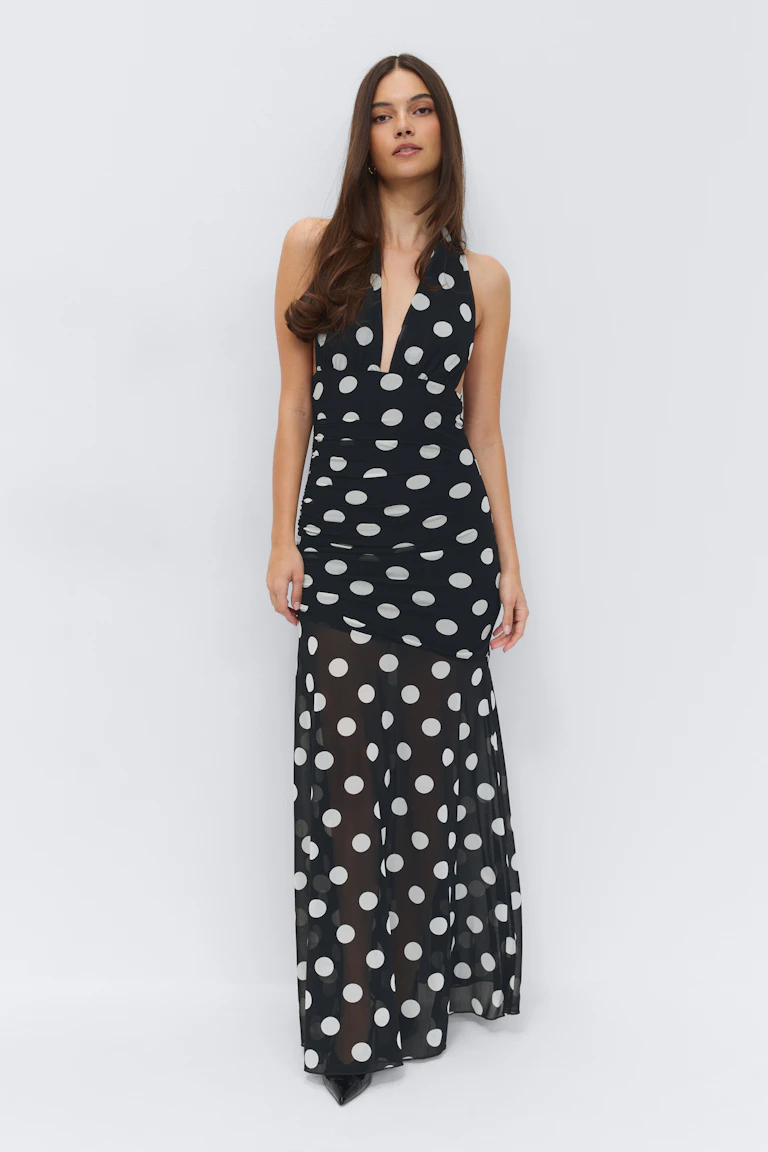 Robe longue noire décolletée avec un profond décolleté en V et des pois blancs, comprenant un corsage ajusté et une jupe transparente à pois, associée à des talons noirs à bouts pointus.