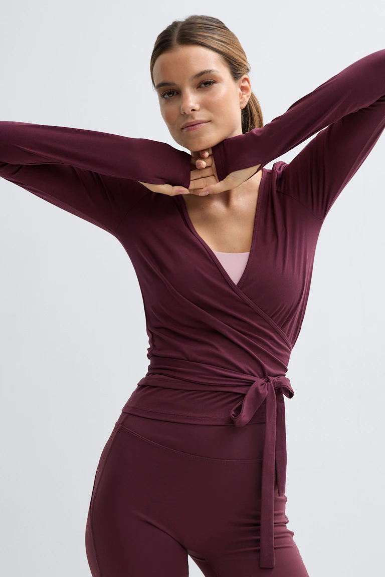 Diep bordeauxrode overslagtop met lange mouwen, duimgaten en een gestrikte taille, over een lichtroze sportbeha, gecombineerd met een bijpassende bordeauxrode legging met hoge taille.