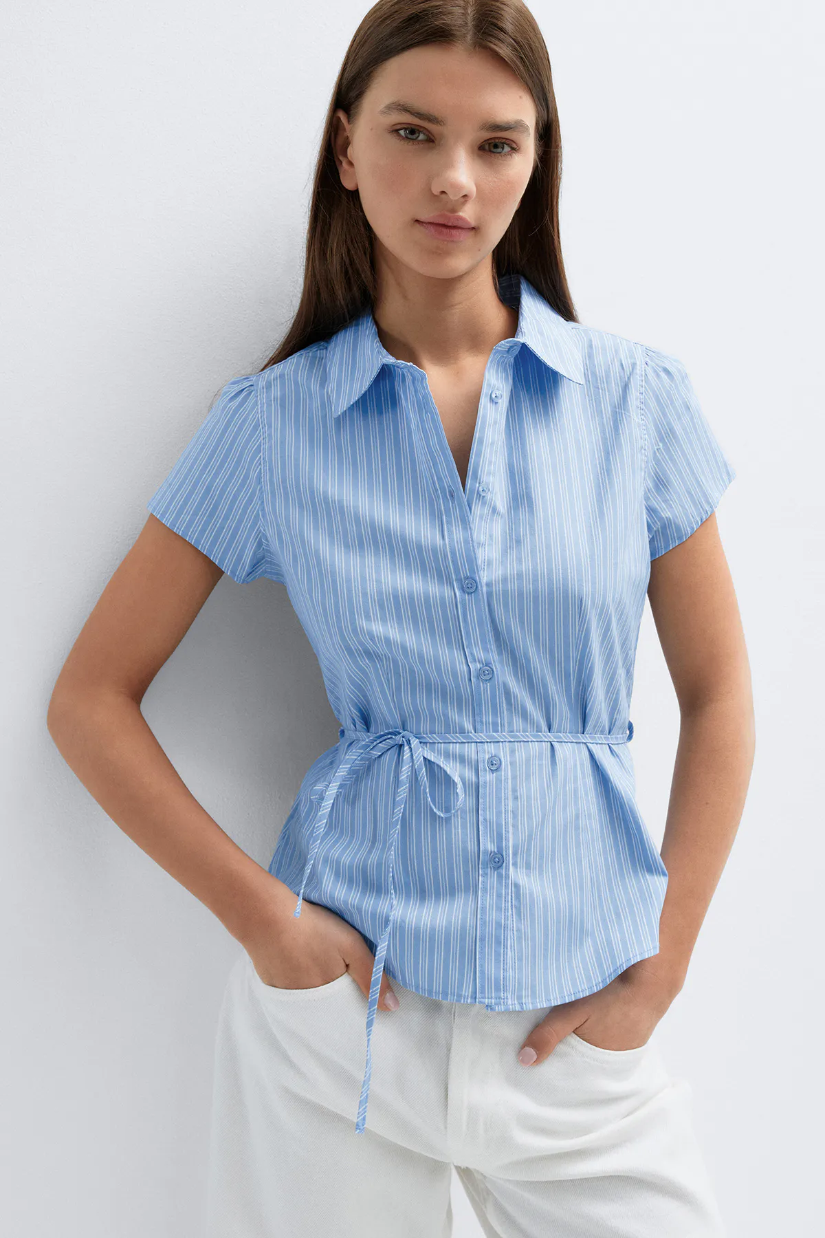Hellblaue Kurzarmbluse mit Knopfleiste, weißen vertikalen Nadelstreifen, spitzem Kragen und einem Gürtel zum Binden in der Taille, kombiniert mit einer weißen Hose.