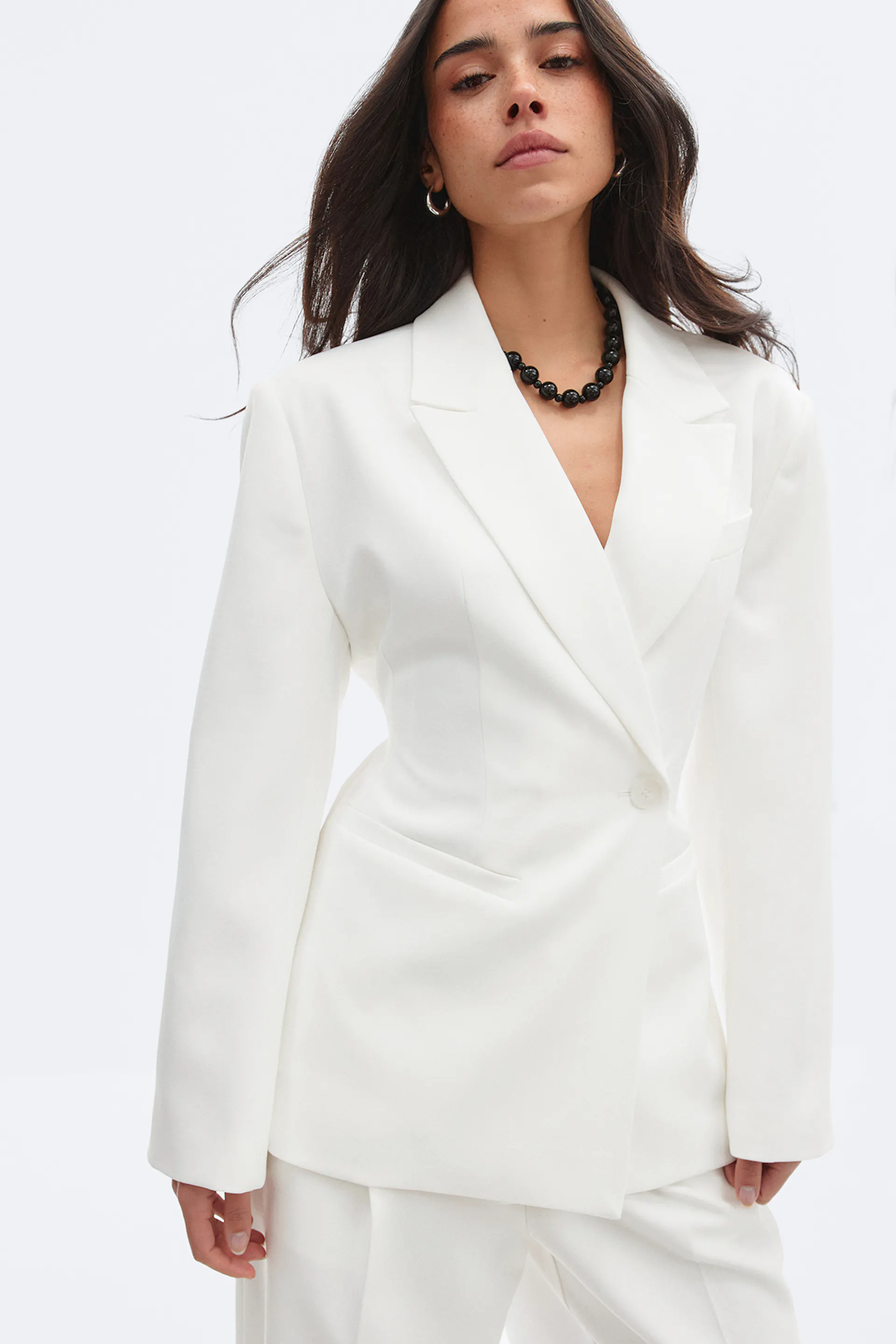 Skræddersyet hvid blazer med en enkelt knap og hakkede revers og matchende hvide bukser, suppleret med en sort perlekæde og små øreringe.