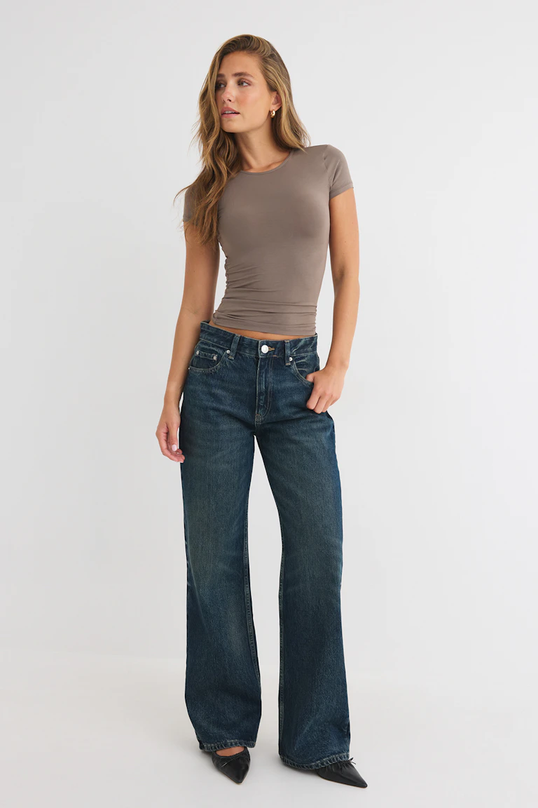 Figurbetontes, taupefarbenes Kurzarm-Oberteil, kombiniert mit einer dunklen Jeans mit weitem Bein und schwarzen spitzen Ballerinas.