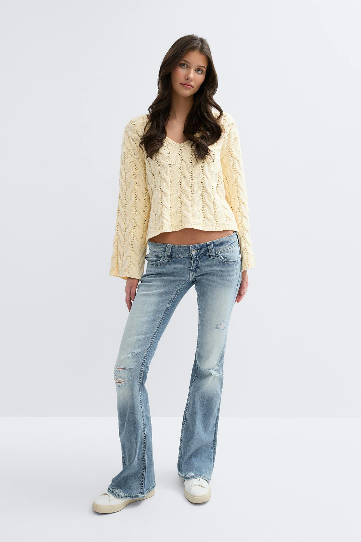 Kermanvärinen kaapelineulottu V-pääntie, vaaleanväriset distressed matalavyötäröiset levenevät farkut ja valkoiset matalat tennarit.