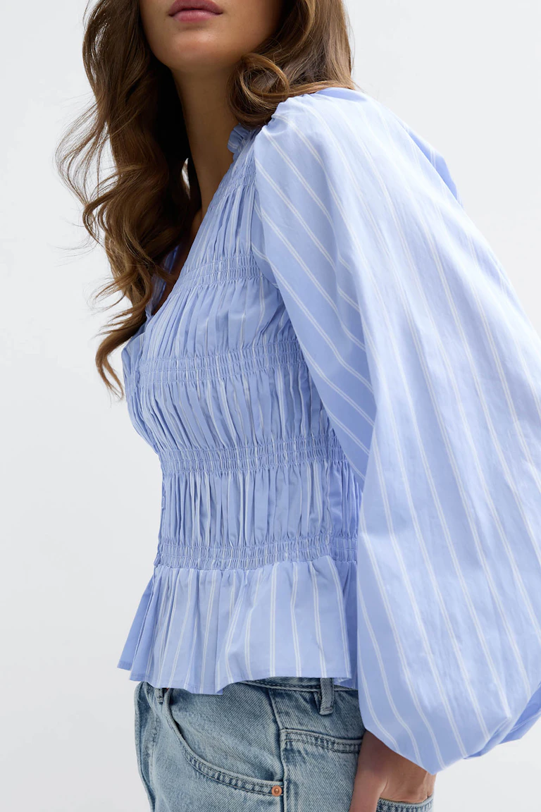 Hellblaue Bluse mit weißen Nadelstreifen, gesmoktem, tailliertem Oberteil, Schößchensaum und voluminösen langen Ärmeln, kombiniert mit hellen Denim-Jeans.