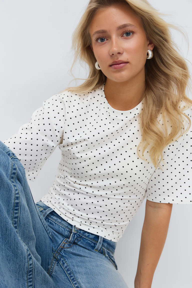 Witte top met korte mouwen en kleine zwarte stippen, gecombineerd met een high-rise jeans en geribde gouden oorbellen.