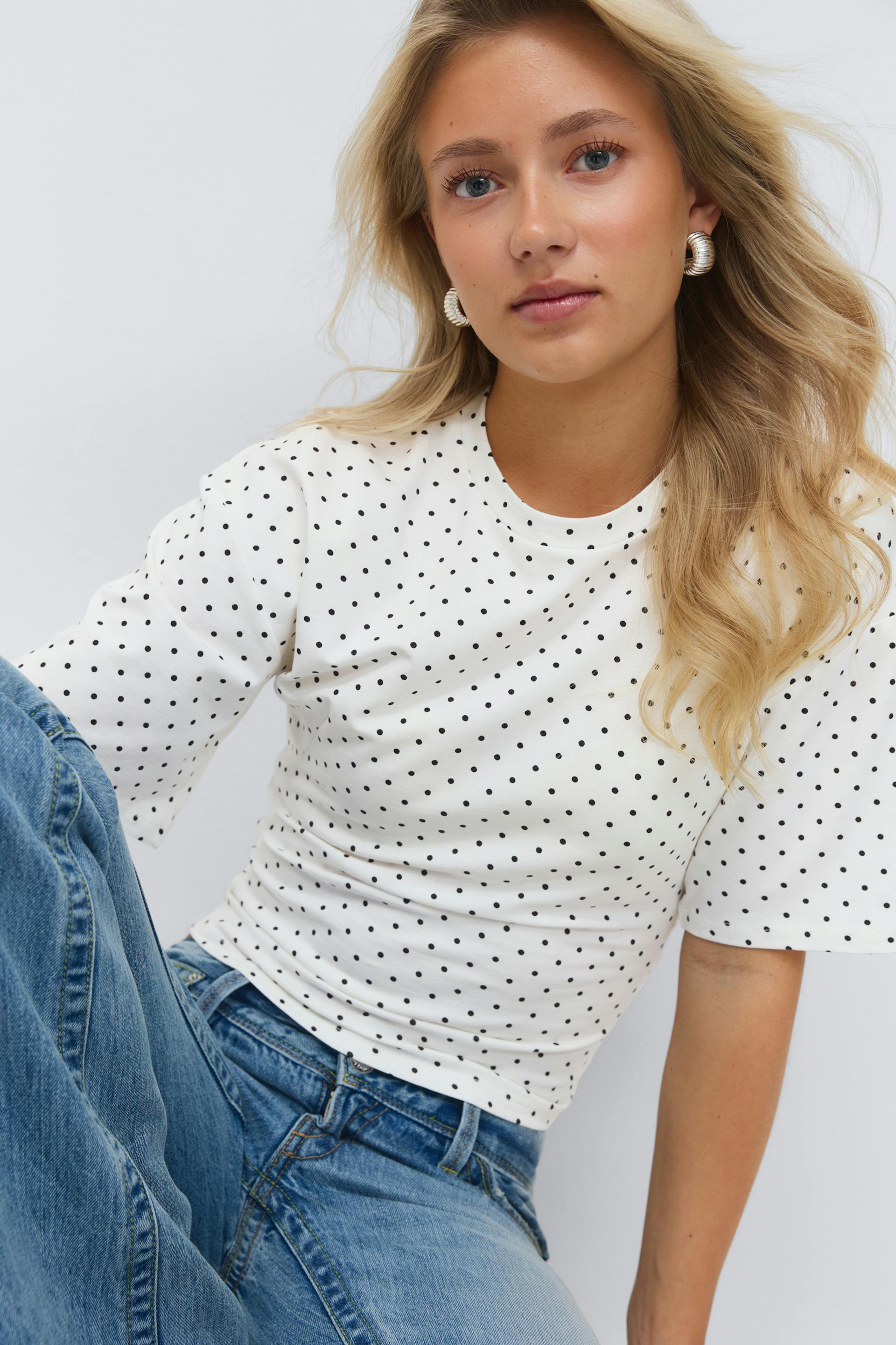 Valkoinen, lyhythihainen toppi, jossa on pieniä mustia pilkkuja, yhdistettynä vaaleisiin korkeavyötäröisiin farkkuihin ja uurrettuihin kultaisiin korvakoruihin.