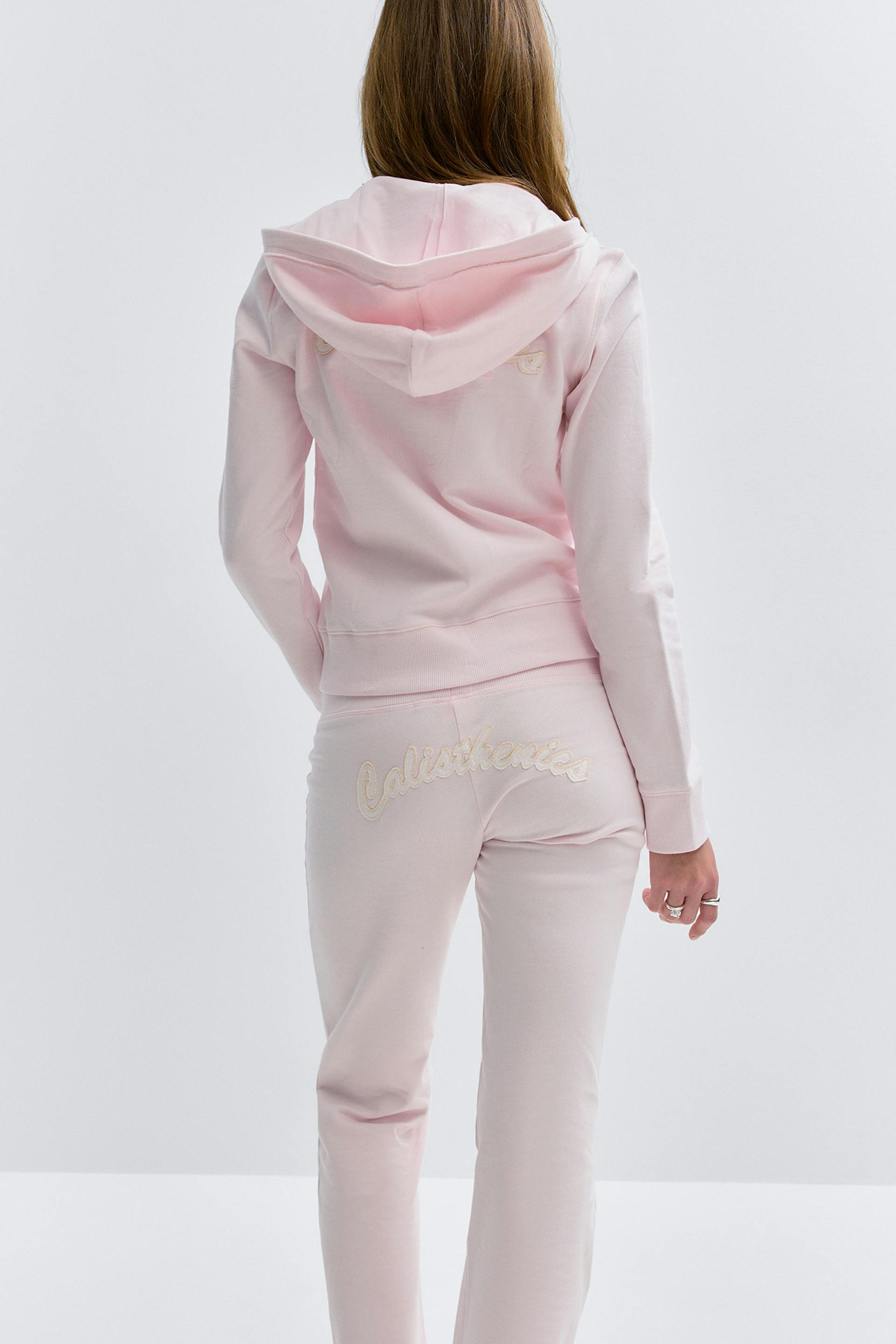 Hellrosa Kapuzensweatshirt mit Rippbündchen an Saum und Ärmeln, kombiniert mit einer passenden hellrosa Jogginghose mit cremefarbenem gesticktem "Calisthenics"-Schriftzug auf dem Rücken.