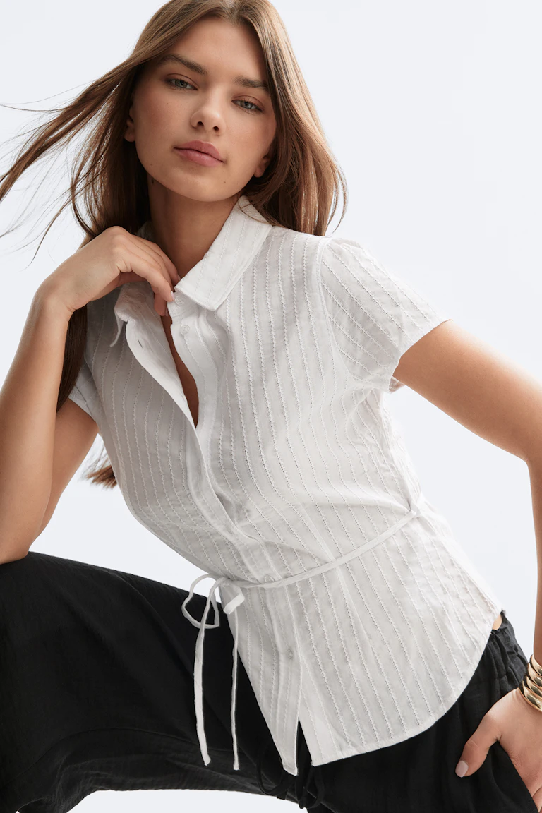 Witte blouse met korte mouwen en knoopsluiting, subtiele verticale plooitjes en zelfknoopsluiting in de taille, gecombineerd met een losse zwarte broek en gestapelde gouden armbanden.