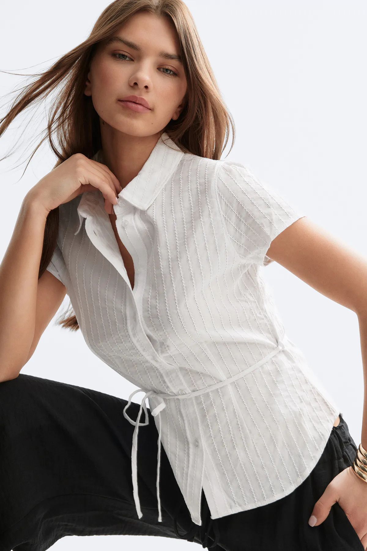 Weiße, kurzärmelige Bluse mit Knopfleiste, dezenten vertikalen Biesenstreifen und einer selbstgebundenen Taille, kombiniert mit einer weiten schwarzen Hose und goldenen Armreifen.