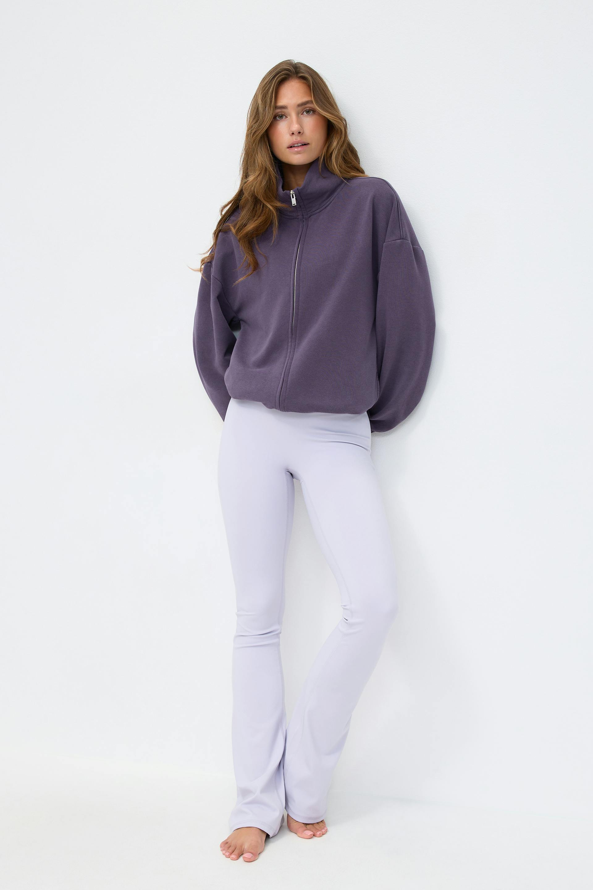 Übergroßes Sweatshirt mit hohem Kragen und Reißverschluss in dunkler Pflaume, kombiniert mit hochtaillierten, lavendelfarbenen Schlagleggings; barfuß.