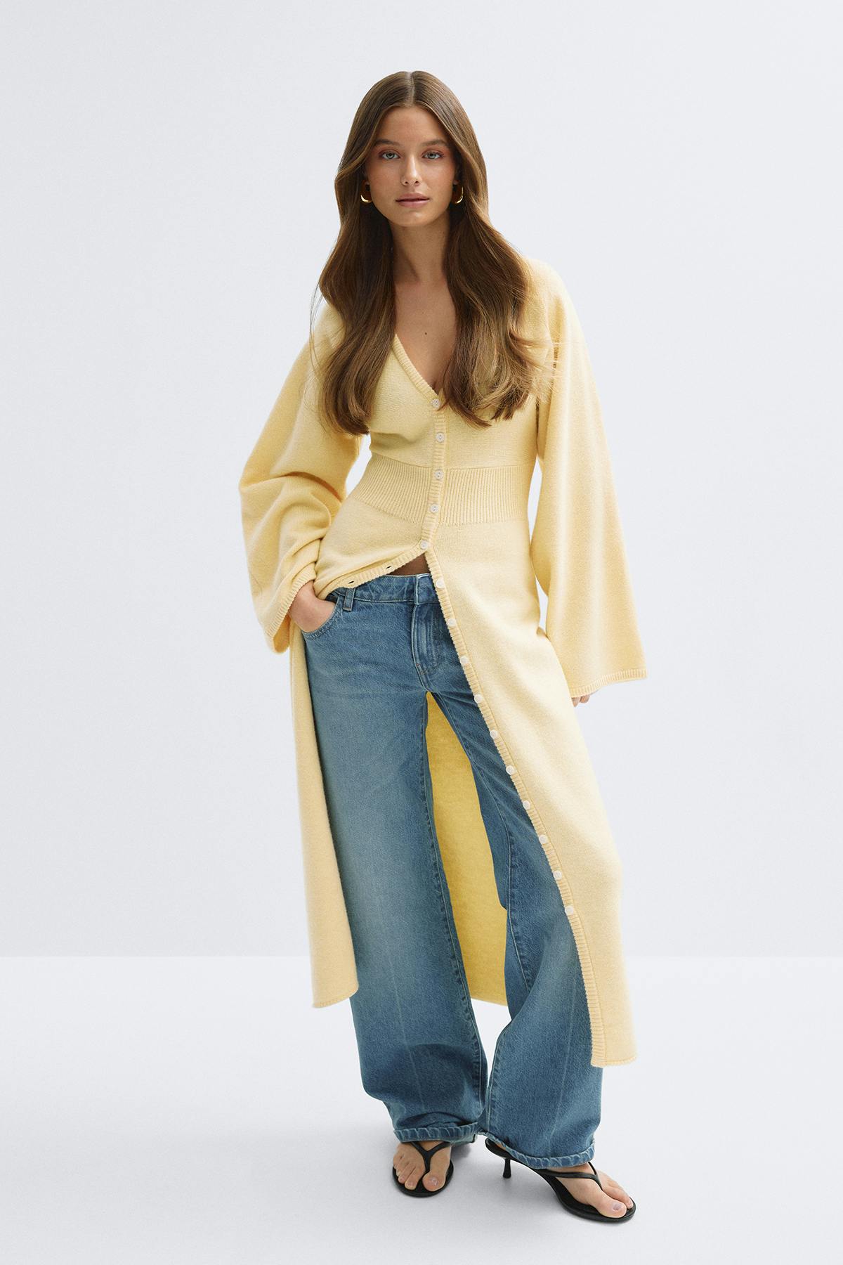 Cardigan long en maille boutonnée jaune pâle avec taille côtelée et larges manches ballon, associé à un jean large bleu clair, des sandales à talons noires et de petites boucles d'oreilles dorées.