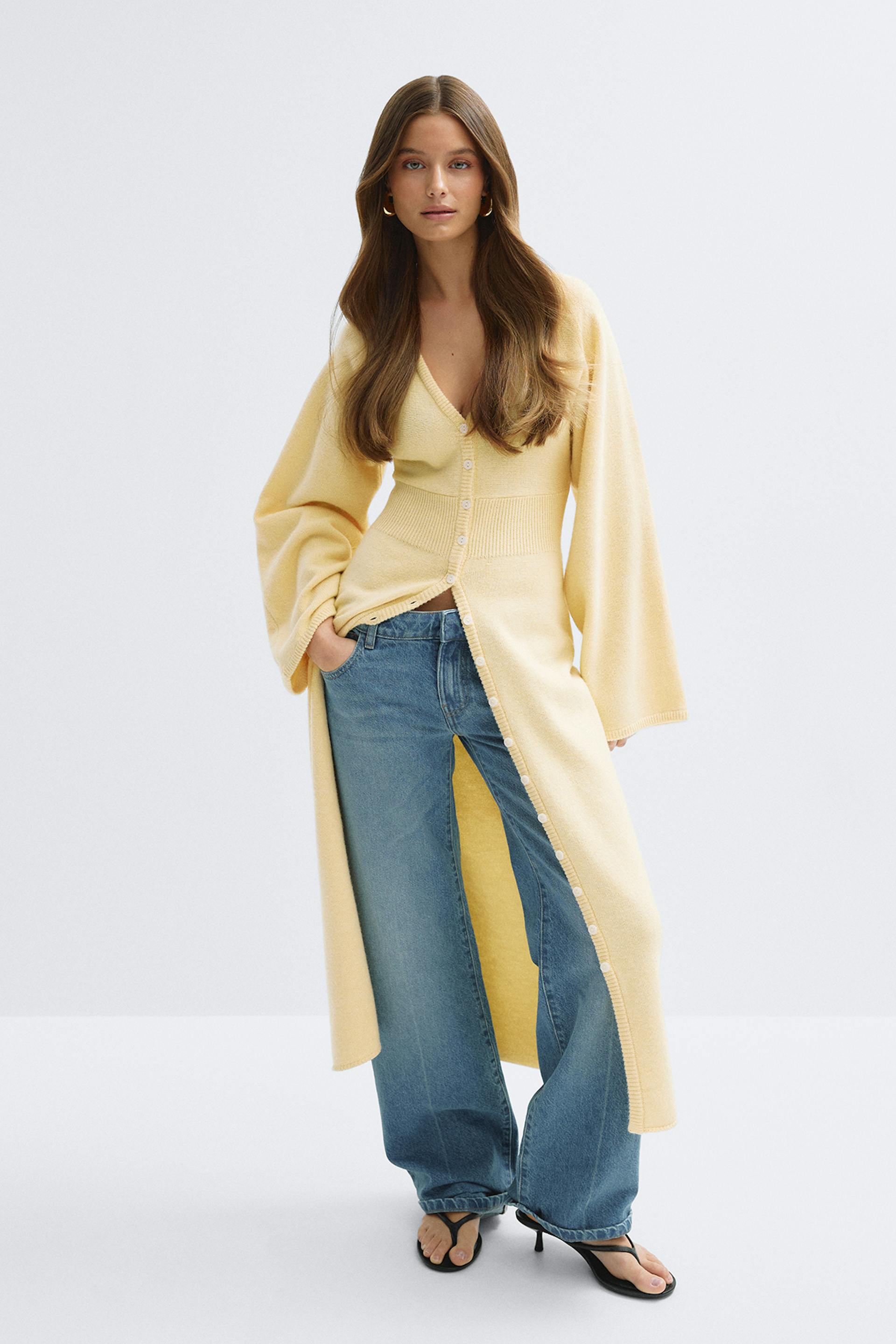 Cardigan long en maille boutonnée jaune pâle avec taille côtelée et larges manches ballon, associé à un jean large bleu clair, des sandales à talons noires et de petites boucles d'oreilles dorées.