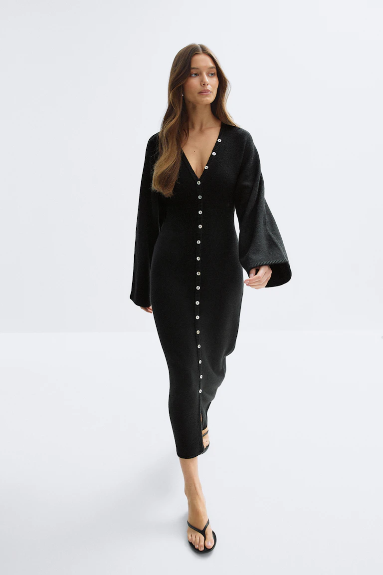 Robe midi noire ajustée en maille côtelée avec col en V, longues manches cloche et rangée centrale de boutons argentés sur une petite fente devant, associée à des sandales thong noires.
