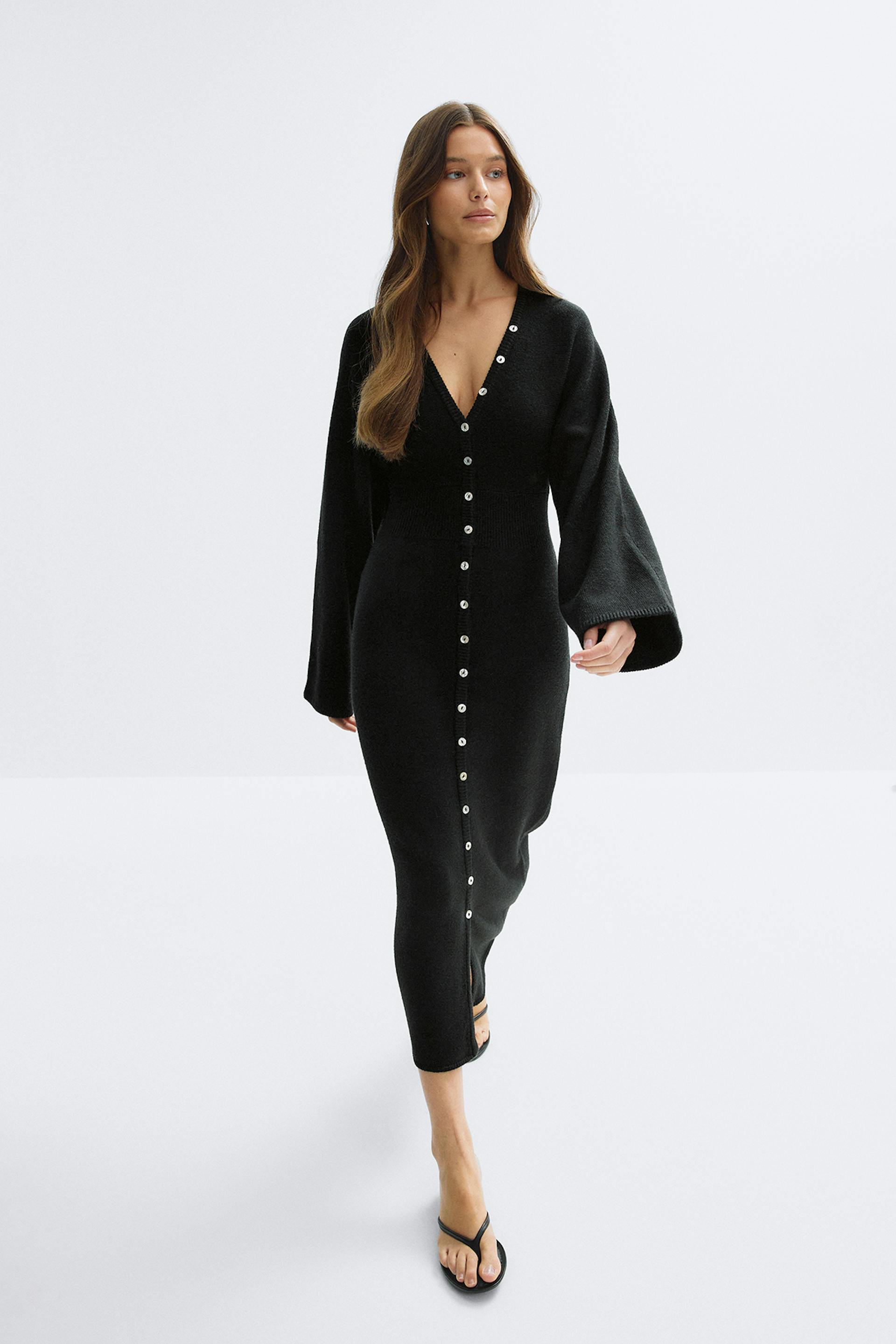 Robe midi noire ajustée en maille côtelée avec col en V, longues manches cloche et rangée centrale de boutons argentés sur une petite fente devant, associée à des sandales thong noires.