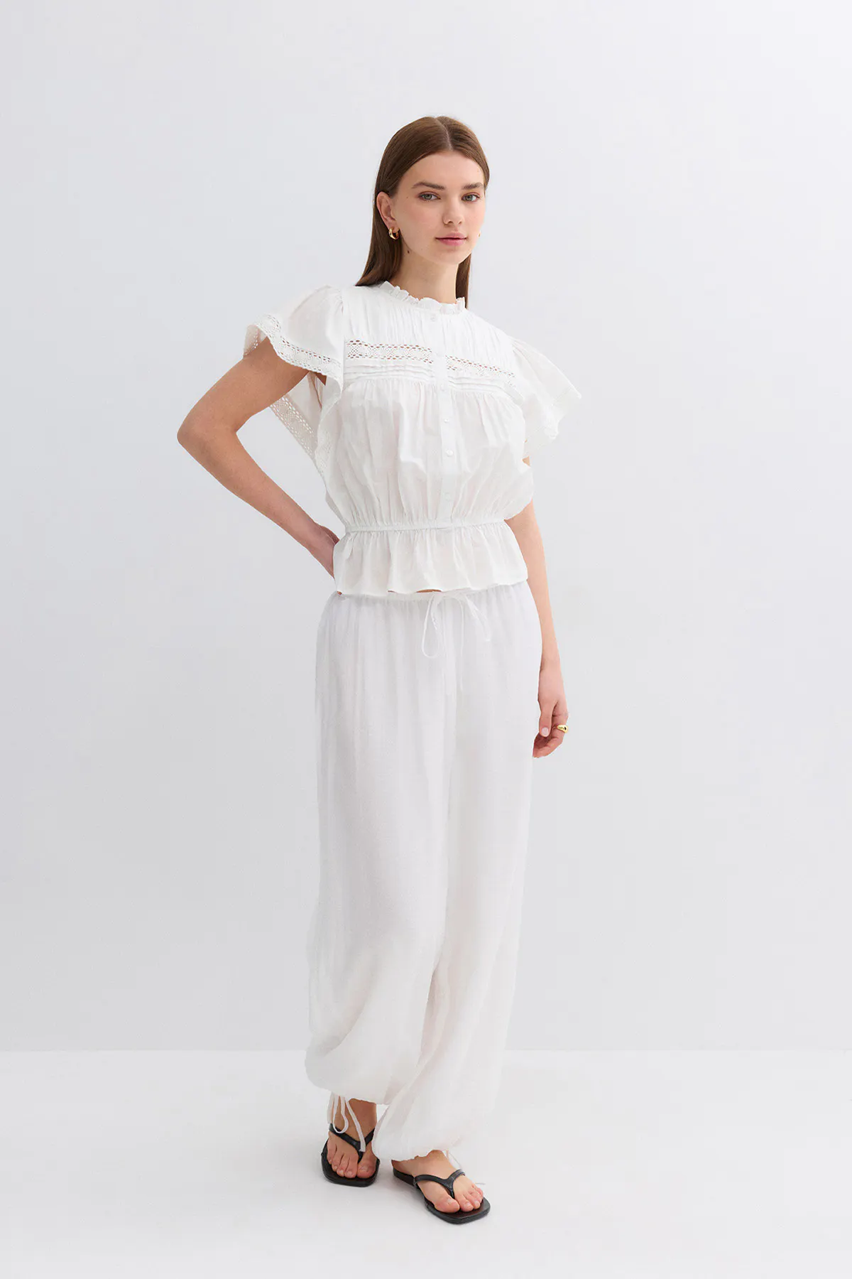 Femme portant un chemisier blanc à manches courtes avec empiècements en dentelle, encolure haute à volants et péplum élastique, associé à un pantalon large à cordon blanc avec chevilles froncées et sandales thong noires ; boucles d'oreilles en or et bague en or.