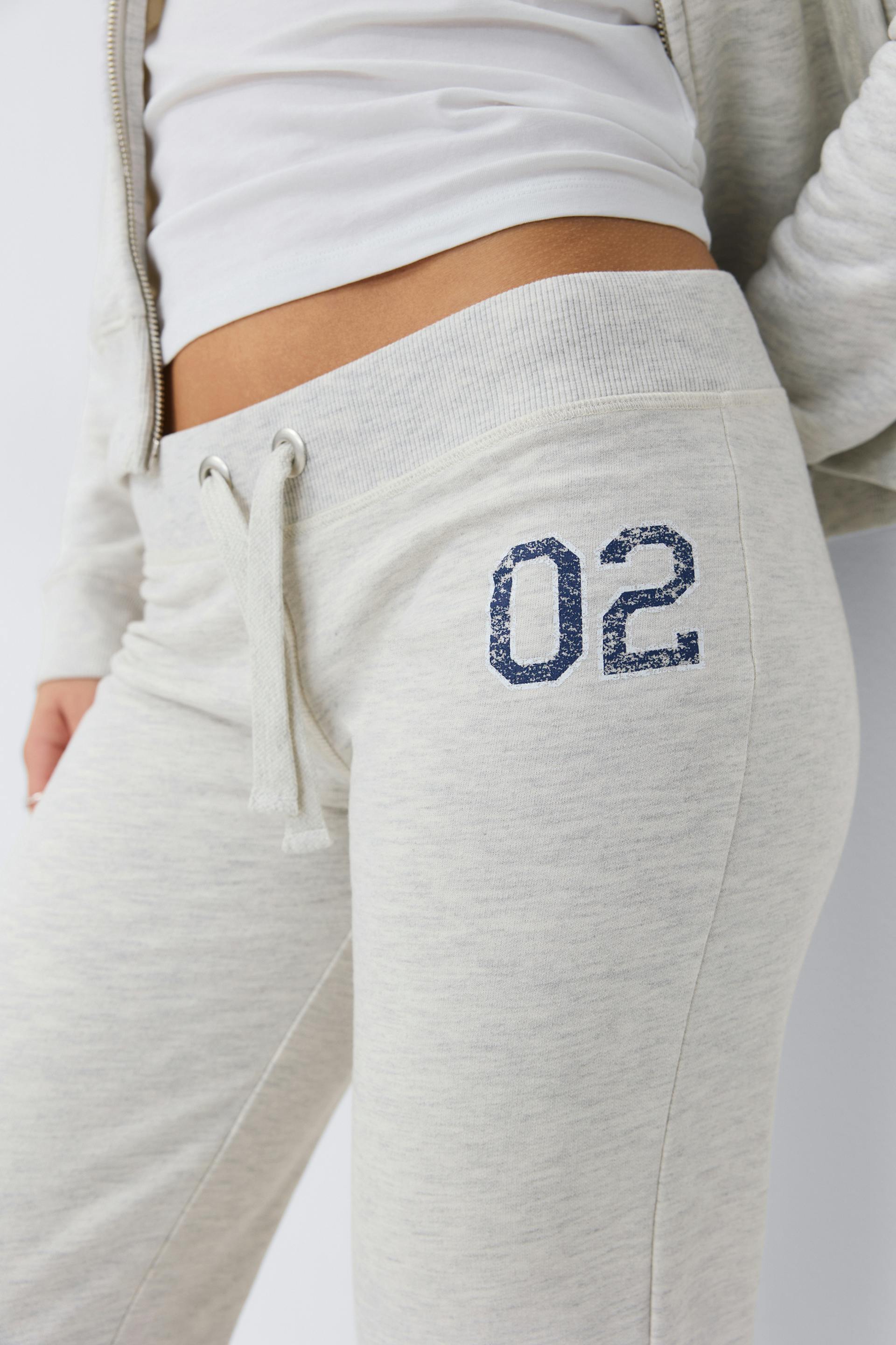 Weißes Kurzarmtank, kombiniert mit einer hellgrauen Jogginghose mit Kordelzug, geripptem Bund und einer marineblauen "02"-Grafik auf der linken Hüfte sowie einem passenden hellgrauen Kapuzenpullover mit Reißverschluss.