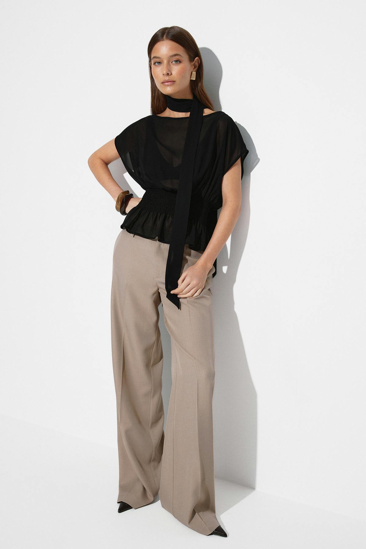 Chemisier noir transparent à manches courtes, à taille smockée et long collier, associé à un pantalon de tailleur beige à jambes larges, à des talons noirs à bouts pointus, à des boucles d'oreilles rectangulaires dorées et à des bracelets bangle en bois.