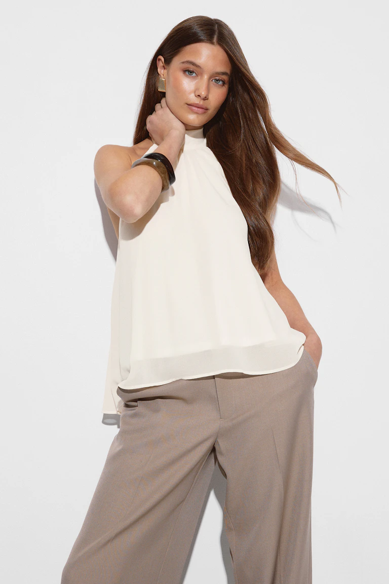 Chemisier en mousseline de soie ivoire sans manches à col haut, associé à un pantalon large taupe, avec des boucles d'oreilles géométriques dorées et un bracelet en écaille foncé.