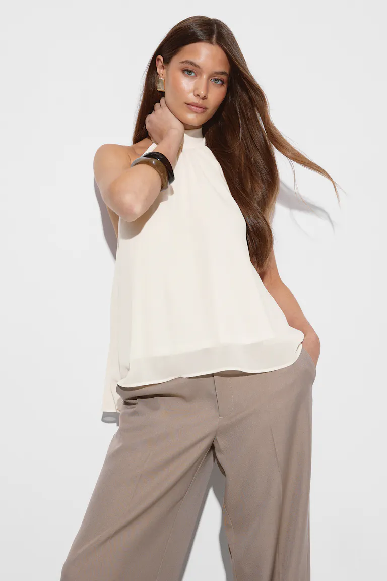 Chemisier en mousseline de soie ivoire sans manches à col haut, associé à un pantalon large taupe, avec des boucles d'oreilles géométriques dorées et un bracelet en écaille foncé.