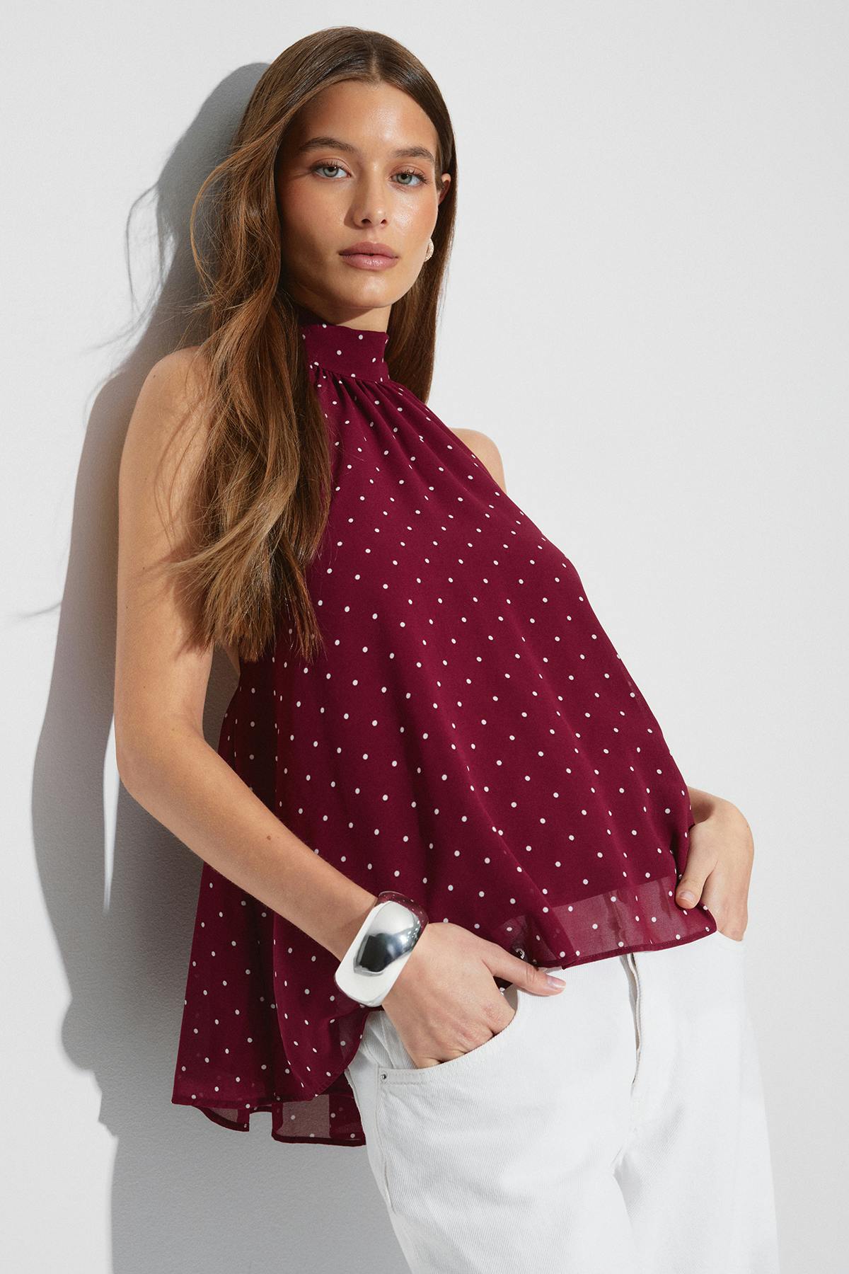Ärmellose burgunderfarbene gepunktete Bluse mit hohem Halsausschnitt und lockerer A-Linien-Silhouette, kombiniert mit einer weißen Hose und einem übergroßen silbernen Armreif.