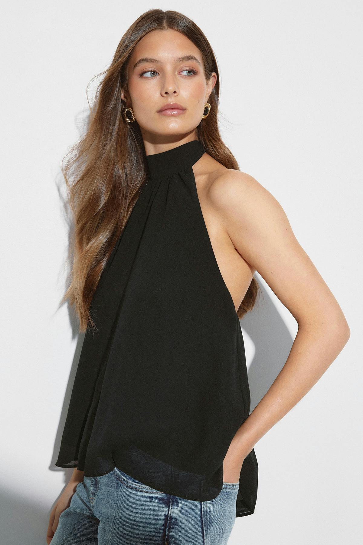 Chemisier noir sans manches à col rond avec un col fantaisie haut et une coupe ample et fluide, associé à un jean bleu délavé et à des boucles d'oreilles dorées texturées.
