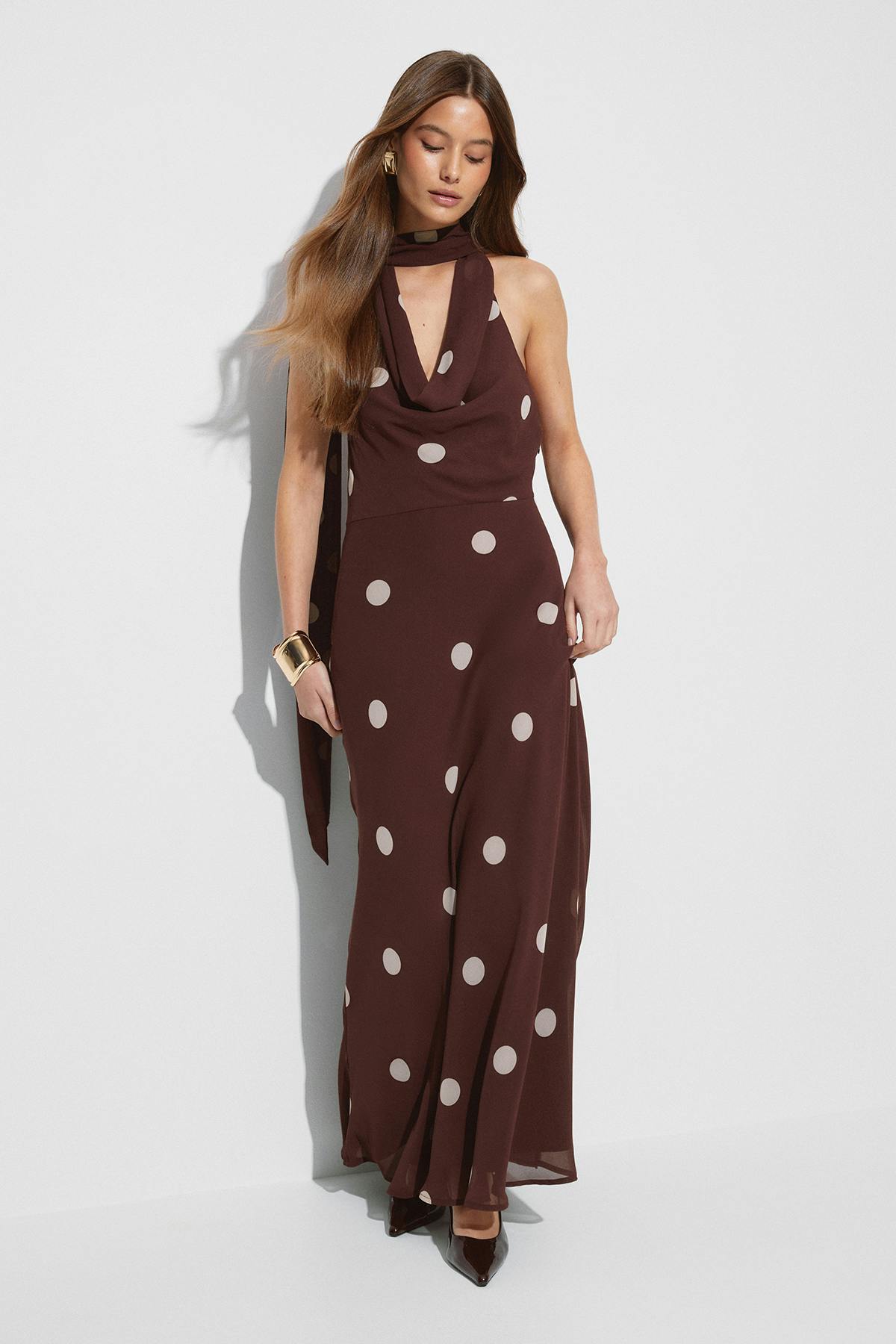 Ermeløs, brun maxikjole med hvite prikker, halterneck og langt, matchende skjerf, sammen med et mansjettarmbånd i gull, geometriske øreringer i gull og spisse, brune lakkpumps.