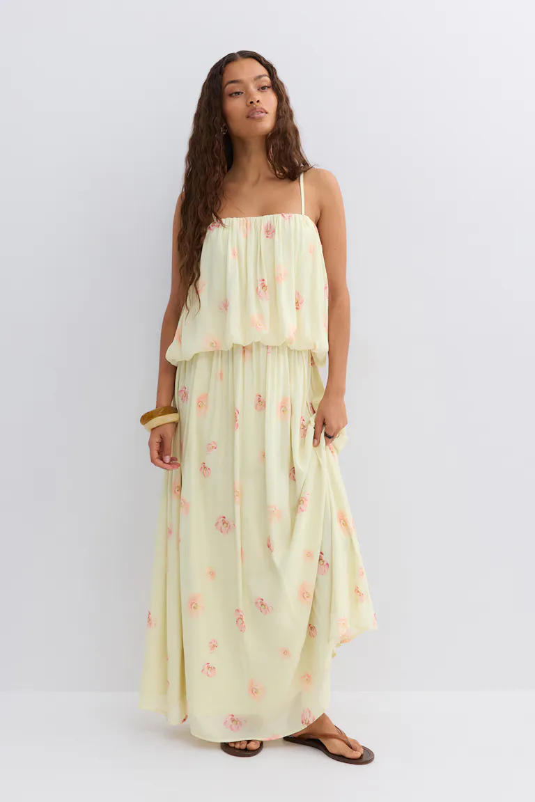 Pale yellow maxi dress with pink floral print, blouson gathered bodice on thin spaghetti straps and a flowing skirt, paired with brown flat sandals and a wooden bangle.