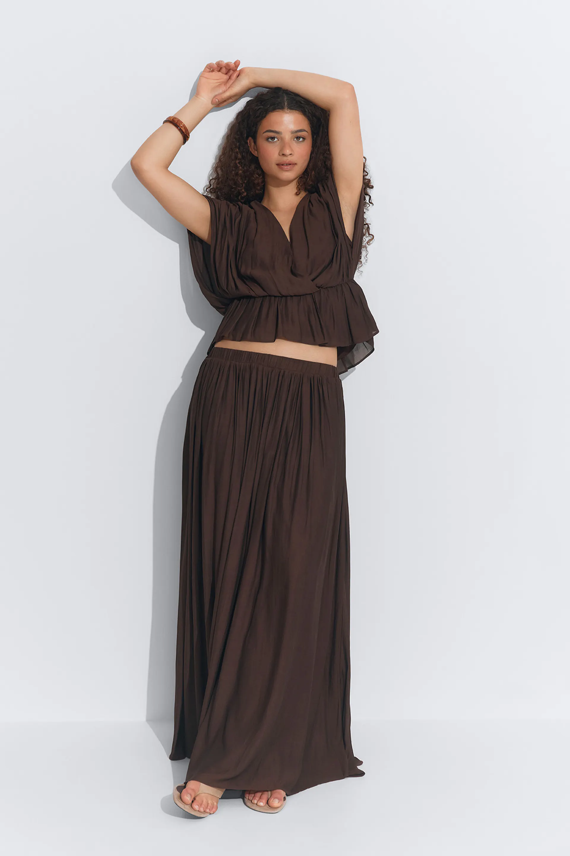 Dunkelbraunes zweiteiliges Set: eine drapierte, kurzärmelige Bluse mit geraffter Taille und ein dazu passender bodenlanger Faltenrock mit hoher Taille; kombiniert mit beigen flachen Sandalen und einem Holzarmreif.