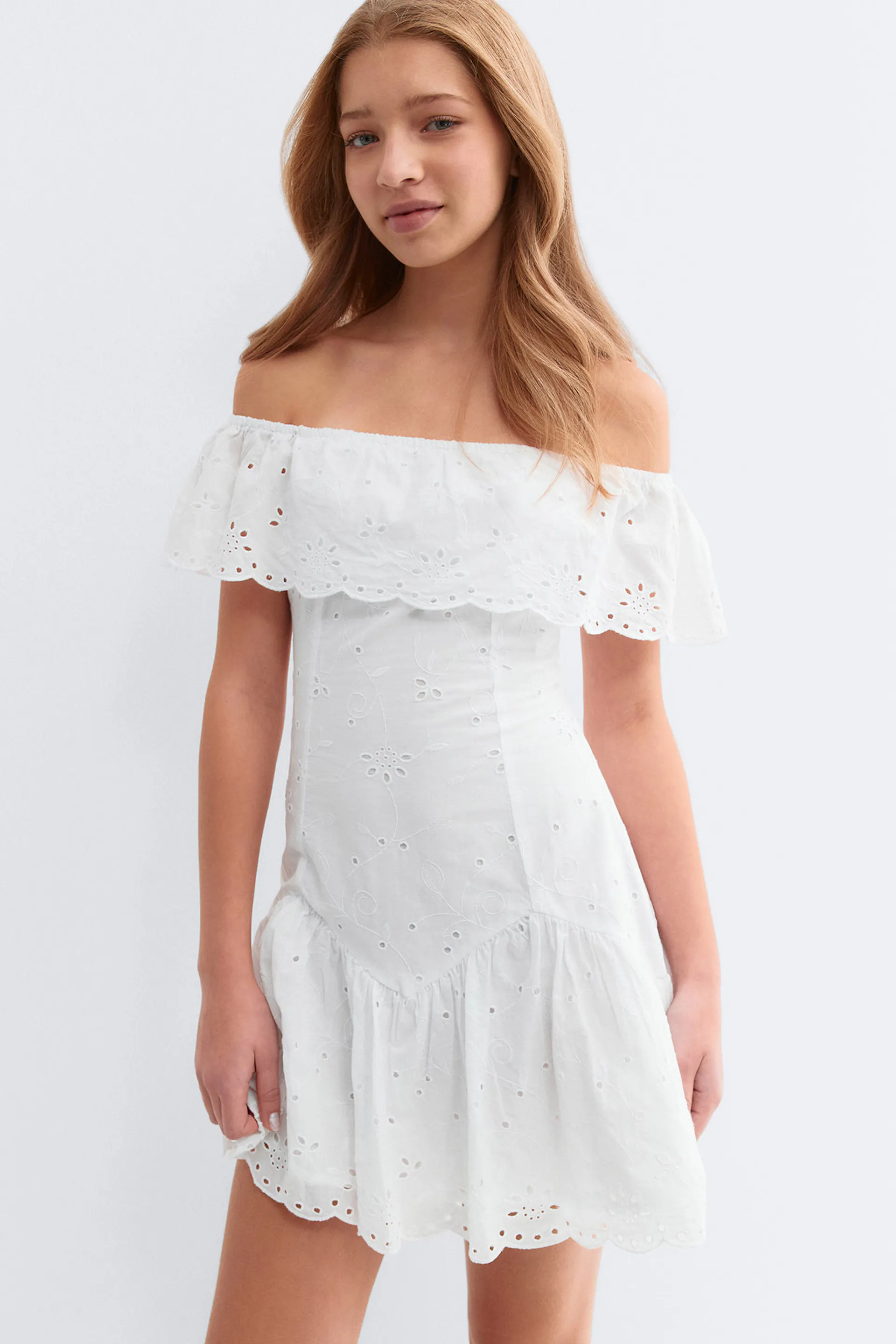 Mini robe blanche à épaules découvertes en broderie anglaise avec encolure à volants, corsage ajusté, jupe évasée à volants et ourlet festonné en œillets.