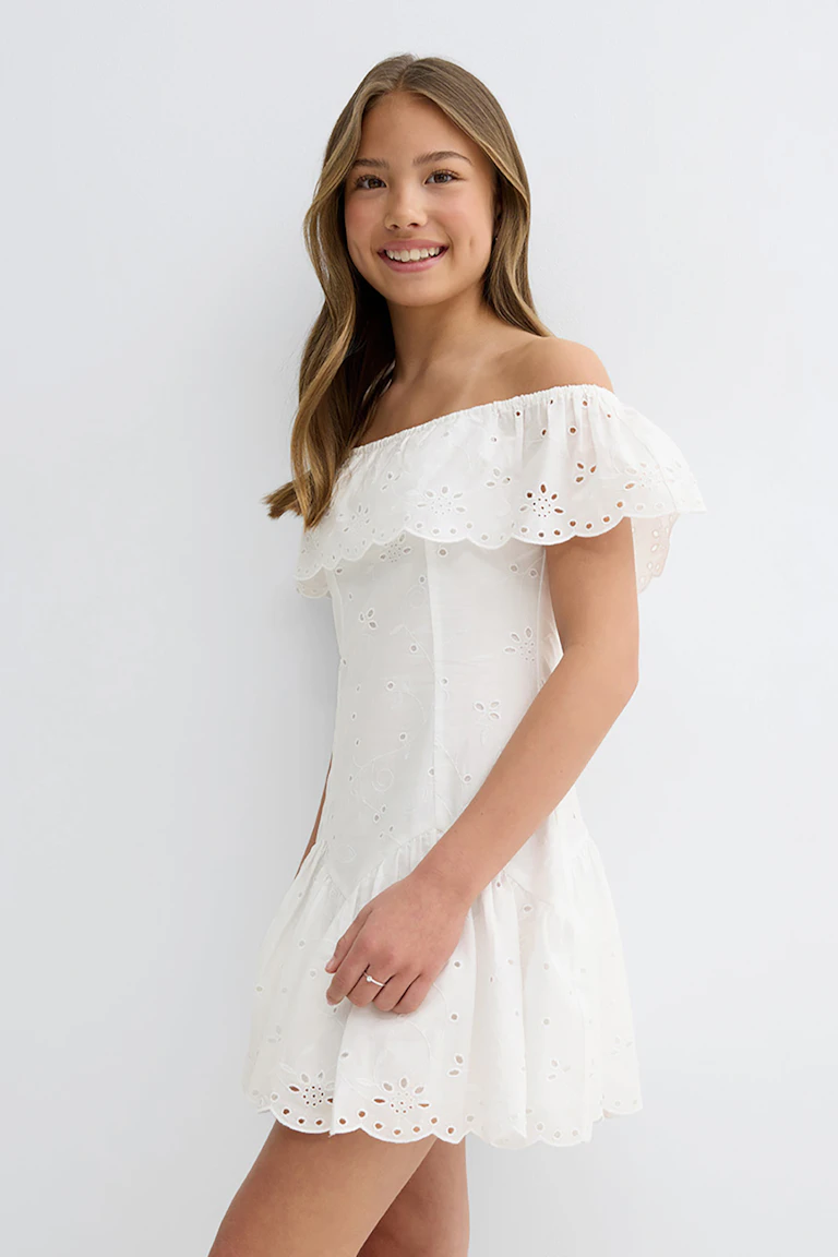 Hvit off-the-shoulder minikjole i broderie anglaise med volangoverlegg i halsen, figursydd overdel, utringet skjørt i etasjeskjørt og kantet øyenkant.