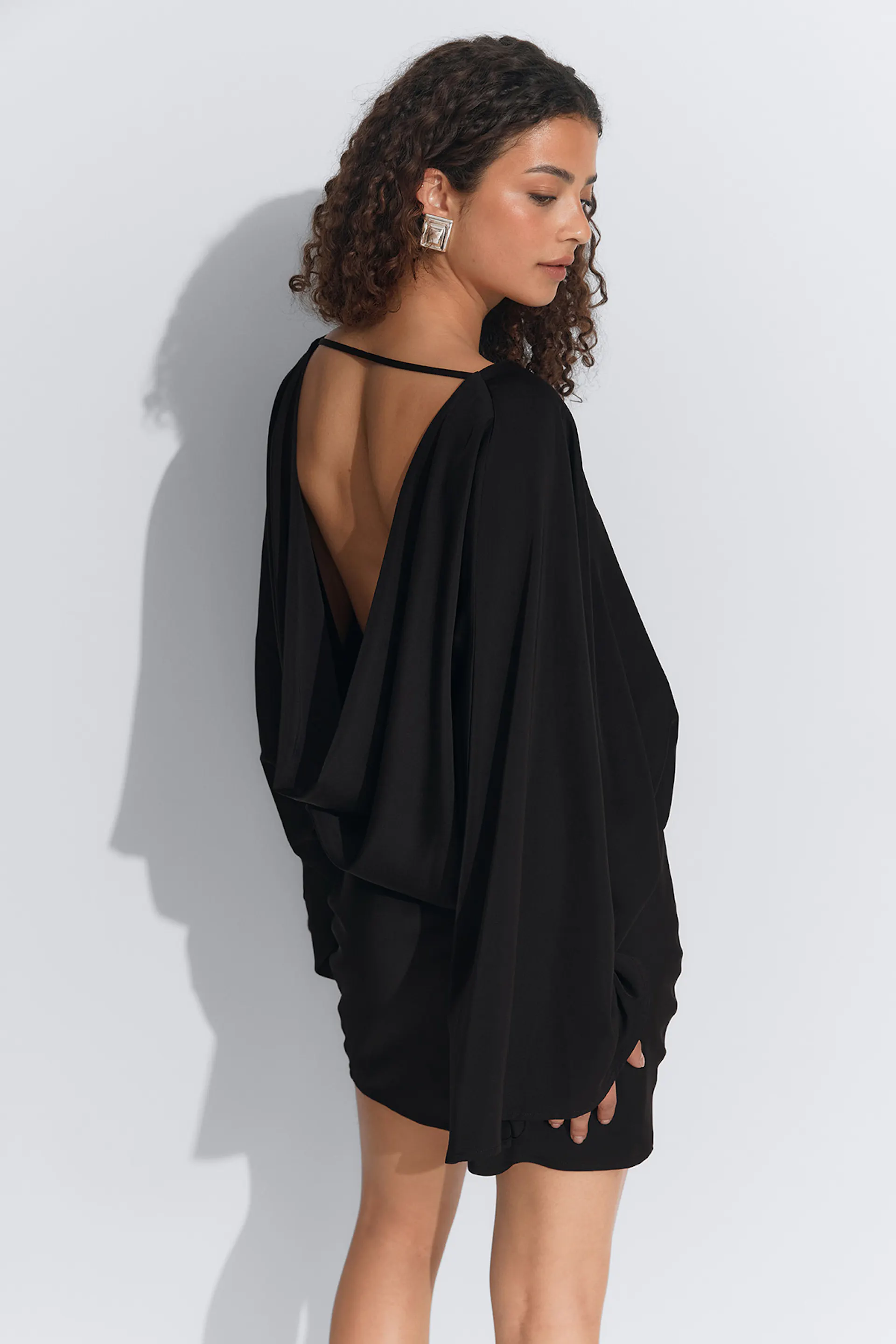 Black draped mini dress with a deep open back secured by a thin upper strap, voluminous batwing sleeves and a fitted above‑knee skirt.