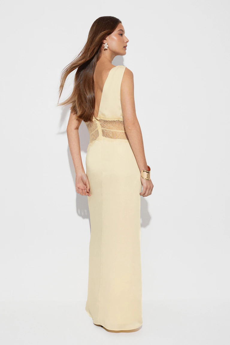 Robe sans manches jaune pâle, longueur du sol, avec un dos en V profond et un panneau de dentelle transparente à la taille, agrémentée de boucles d'oreilles florales et d'un bracelet manchette doré.
