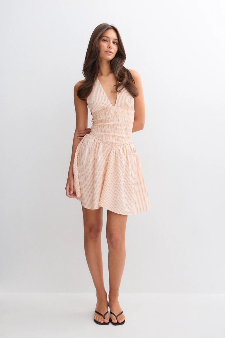 Mini robe dos nu rayée pêche et blanc avec un décolleté en V plongeant, une taille ajustée smockée et une jupe trapèze froncée, associée à des sandales thong noires.
