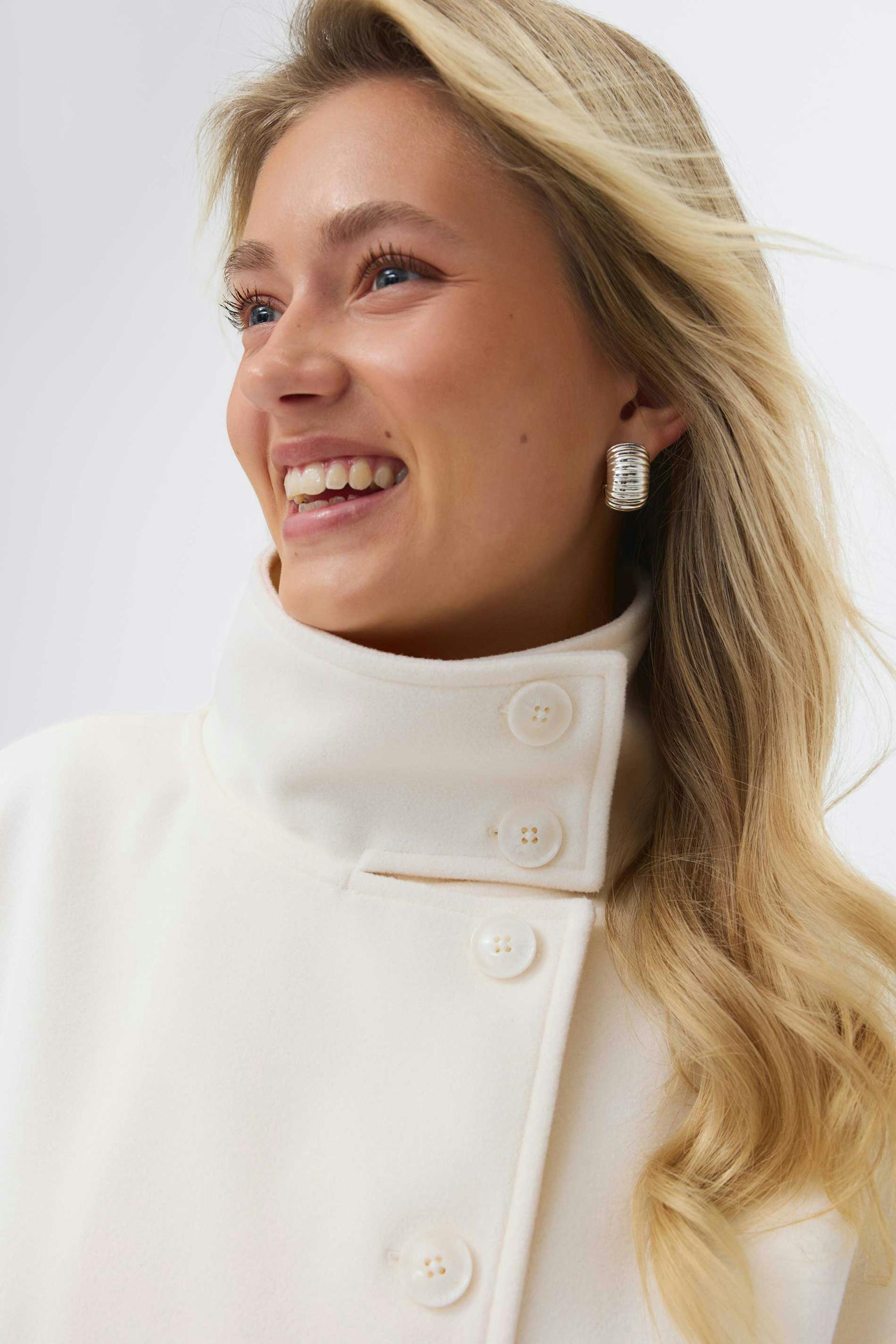Ivory high‑collar wool coat with large round buttons, paired with silver ribbed hoop earrings.